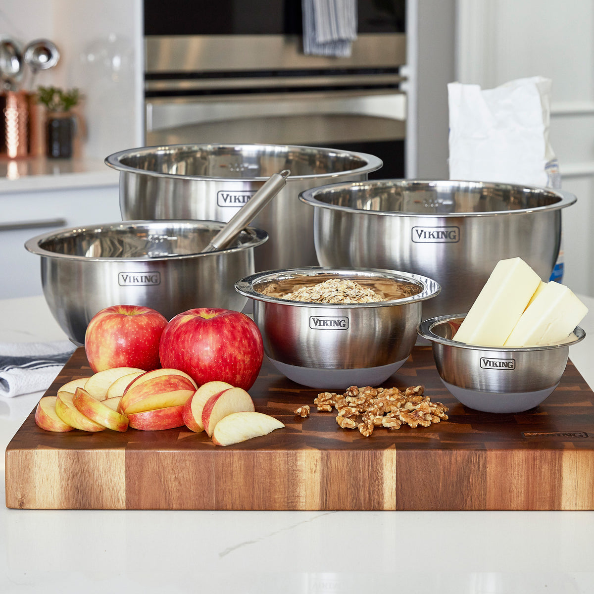 The Viking 10-Piece Stainless Steel Mixing Bowl Set with Lids by Viking Culinary Products sits on a kitchen counter, filled with baking ingredients; sliced apples and walnuts are neatly arranged on a cutting board for compact storage.
