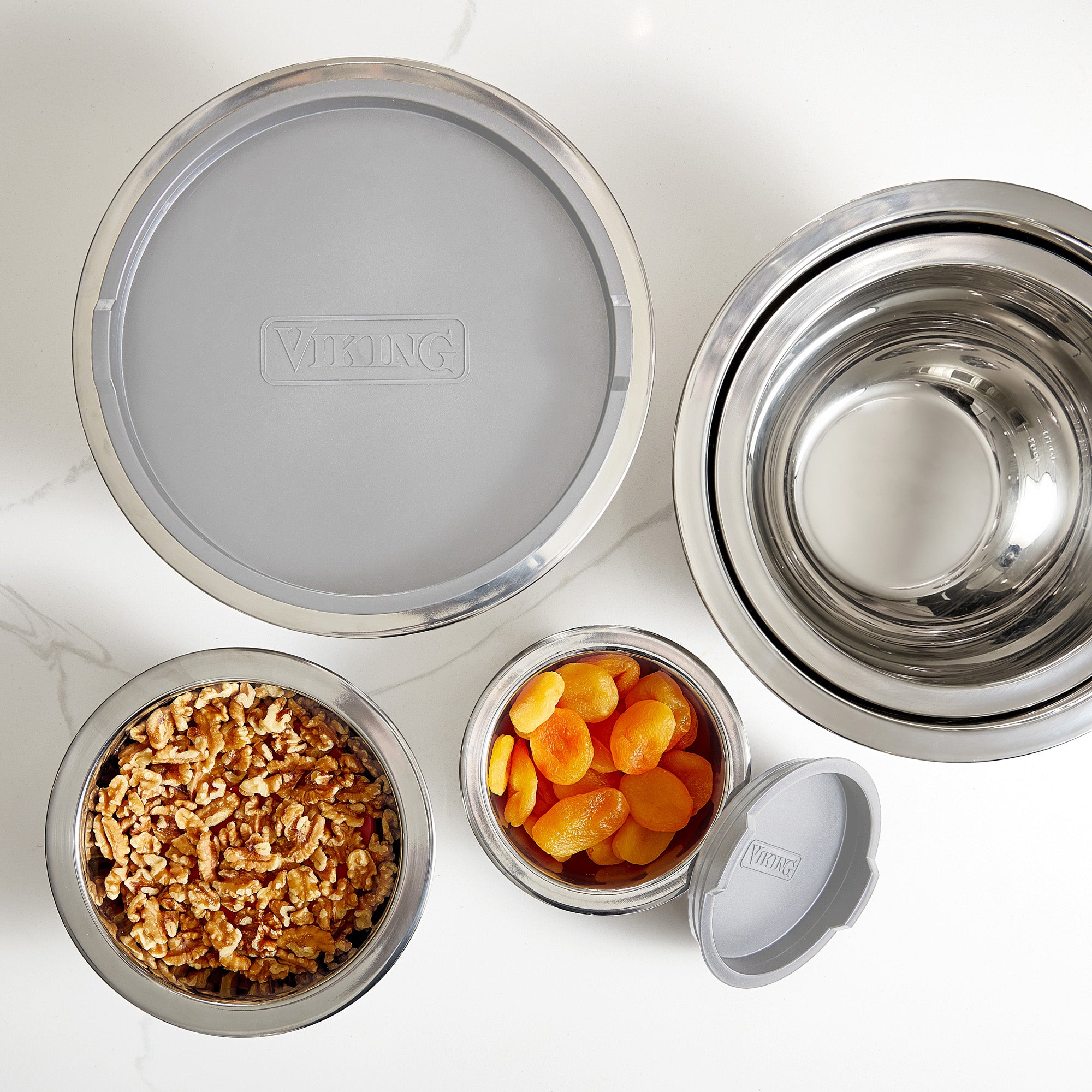 The Viking Culinary Products 10-Piece Stainless Steel Mixing Bowl Set with Lids, Gray, features bowls with silicone bases, shown on white marble—some filled with walnuts and dried apricots, others nested neatly next to a large lidded bowl.