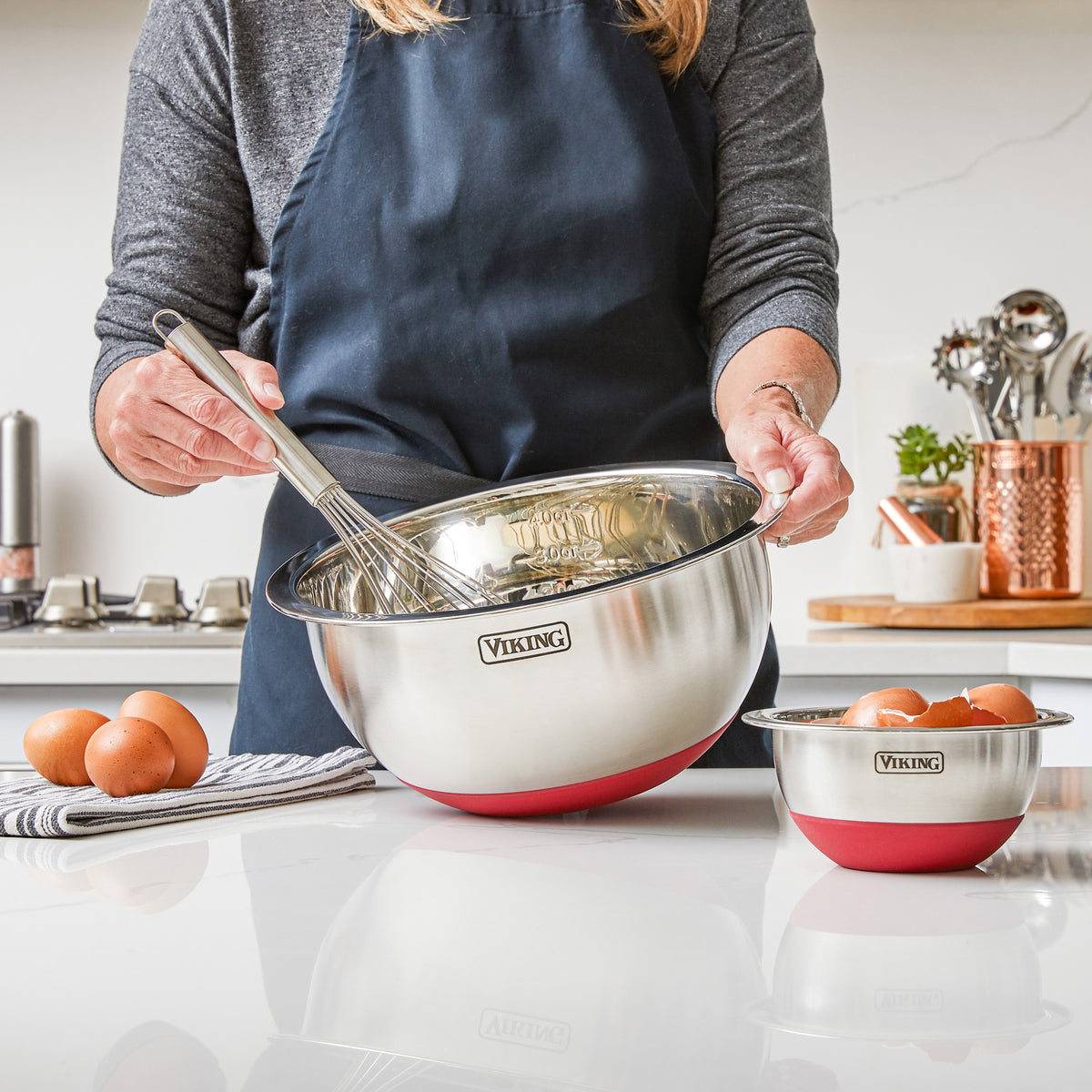 Wearing a navy apron, a person whisks eggs in a stainless steel mixing bowl with silicone base from the Viking 10-Piece Stainless Steel Mixing Bowl Set with Lids by Viking Culinary Products; another bowl and utensils are nearby.