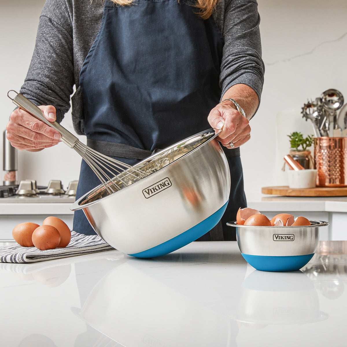 In a kitchen, someone in a navy apron mixes ingredients in a large Viking 10-Piece Stainless Steel Mixing Bowl with Lid (Teal) by Viking Culinary Products. Nearby, a smaller matching bowl holds eggs; both have teal bases and airtight lids for compact storage.