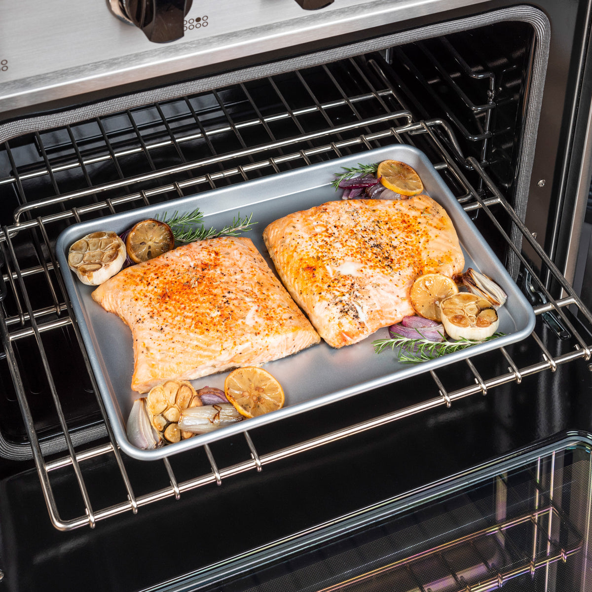 Salmon fillets seasoned with herbs bake on the Viking 15-Inch Aluminized Nonstick Baking Sheet by Viking Culinary Products, surrounded by lemon slices, garlic halves, shallots, and rosemary sprigs.