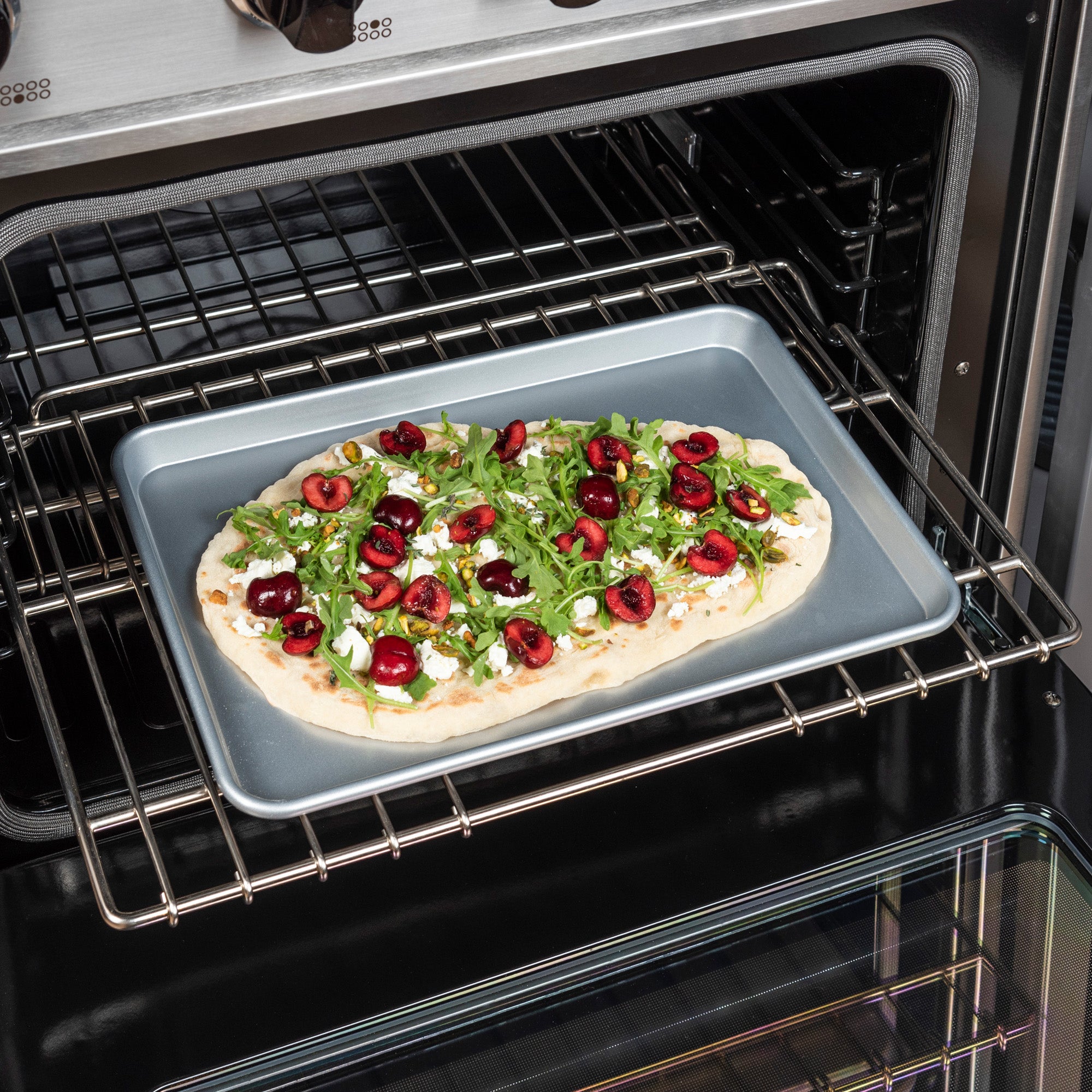 A flatbread with arugula, sliced cherries, and white cheese rests on a Viking Culinary Products Viking 3-Piece Nonstick Aluminized Steel Baking Sheet inside an oven, ready to bake.