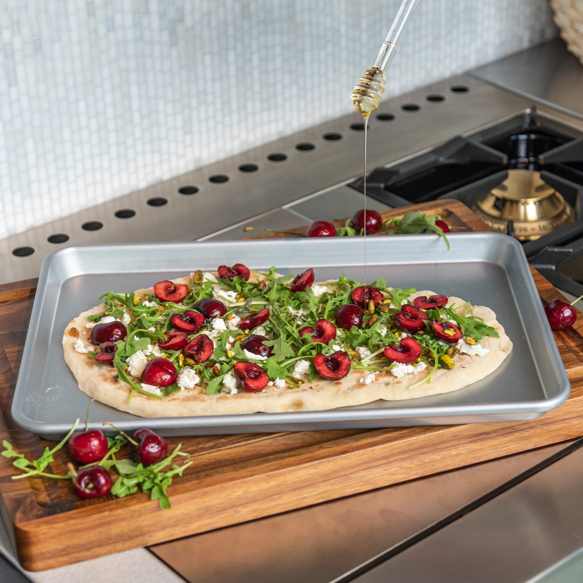 A flatbread with cherries, arugula, pistachios, and cheese sits on a Viking 15-Inch Aluminized Nonstick Baking Sheet by Viking Culinary Products as honey is drizzled on top, all displayed on a kitchen countertop.