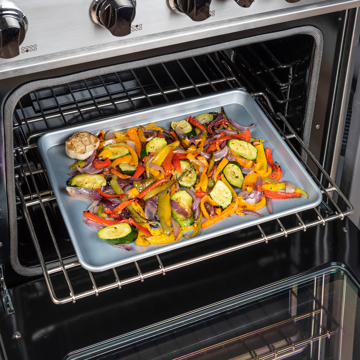 A Viking 17-Inch Aluminized Nonstick Baking Sheet by Viking Culinary Products, filled with sliced bell peppers, zucchini, red onions, and halved garlic, sits on the oven’s middle rack roasting as the open door displays the vibrant vegetables inside.