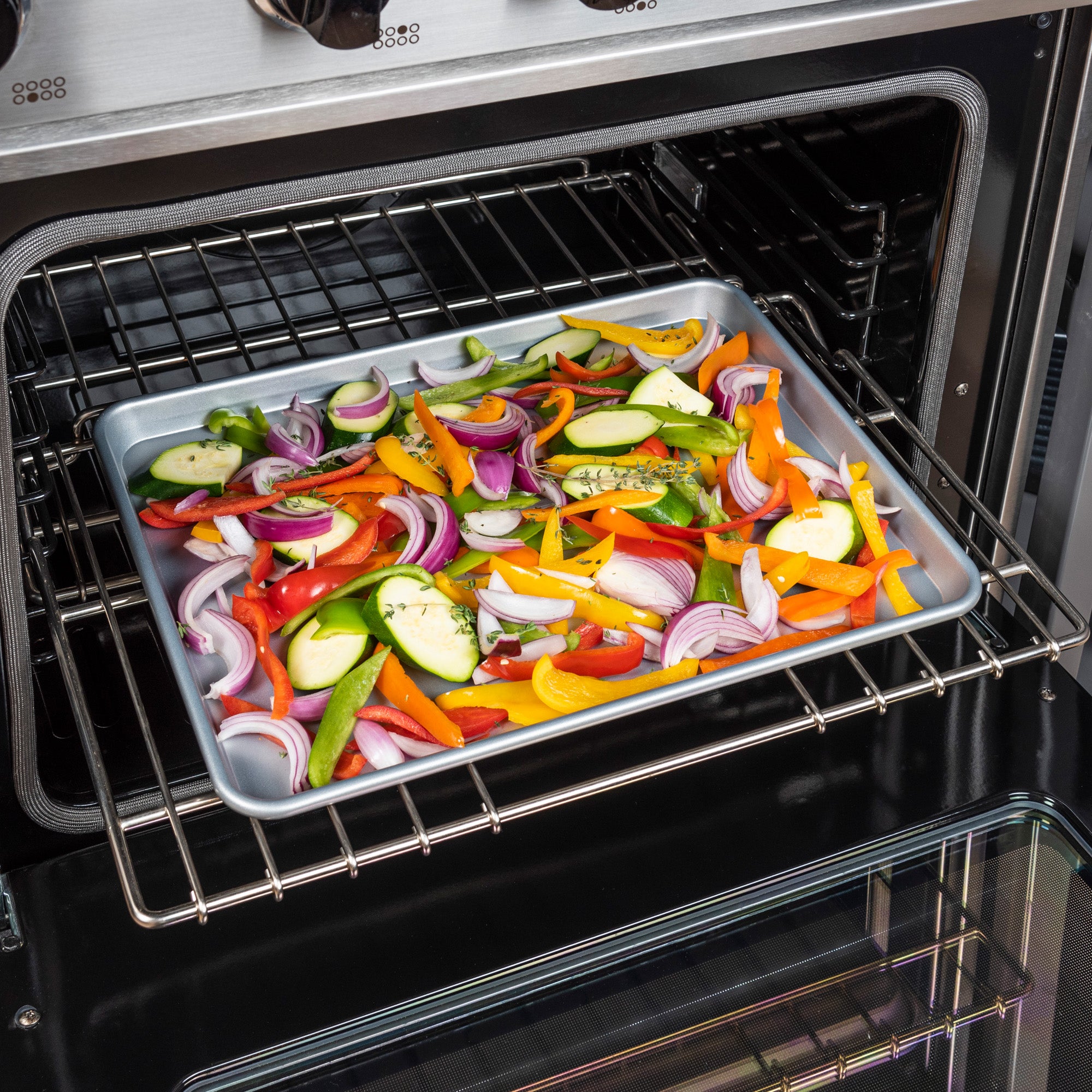 The Viking 3-Piece Nonstick Aluminized Steel Baking Sheet Set by Viking Culinary Products holds sliced zucchini, red onions, and bell peppers, ready for roasting on the oven rack.