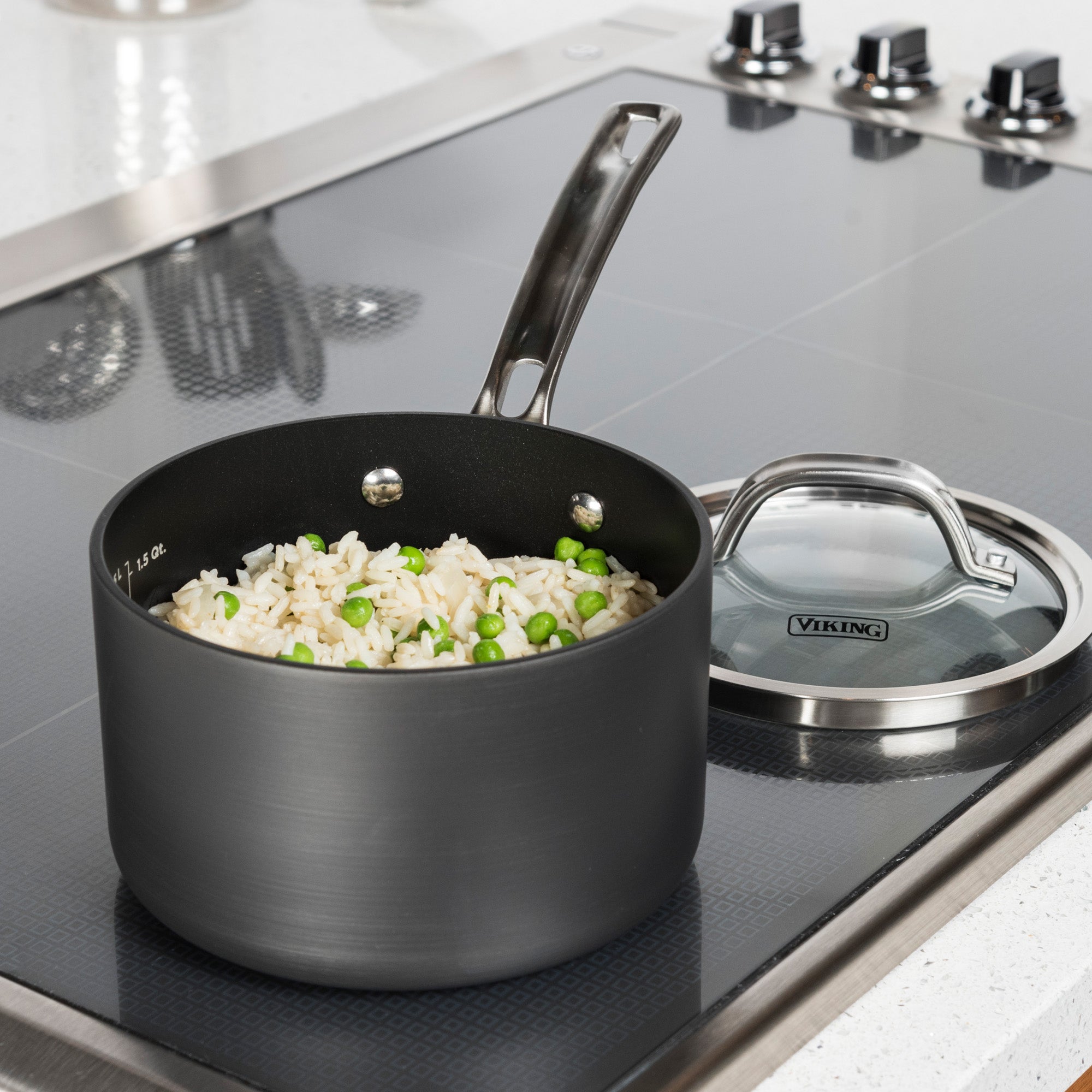 A Viking Hard Anodized Nonstick 2-Quart Sauce Pan with Glass Lid from Viking Culinary Products, containing cooked rice and peas, sits on a stove with four burner knobs beside a white countertop.