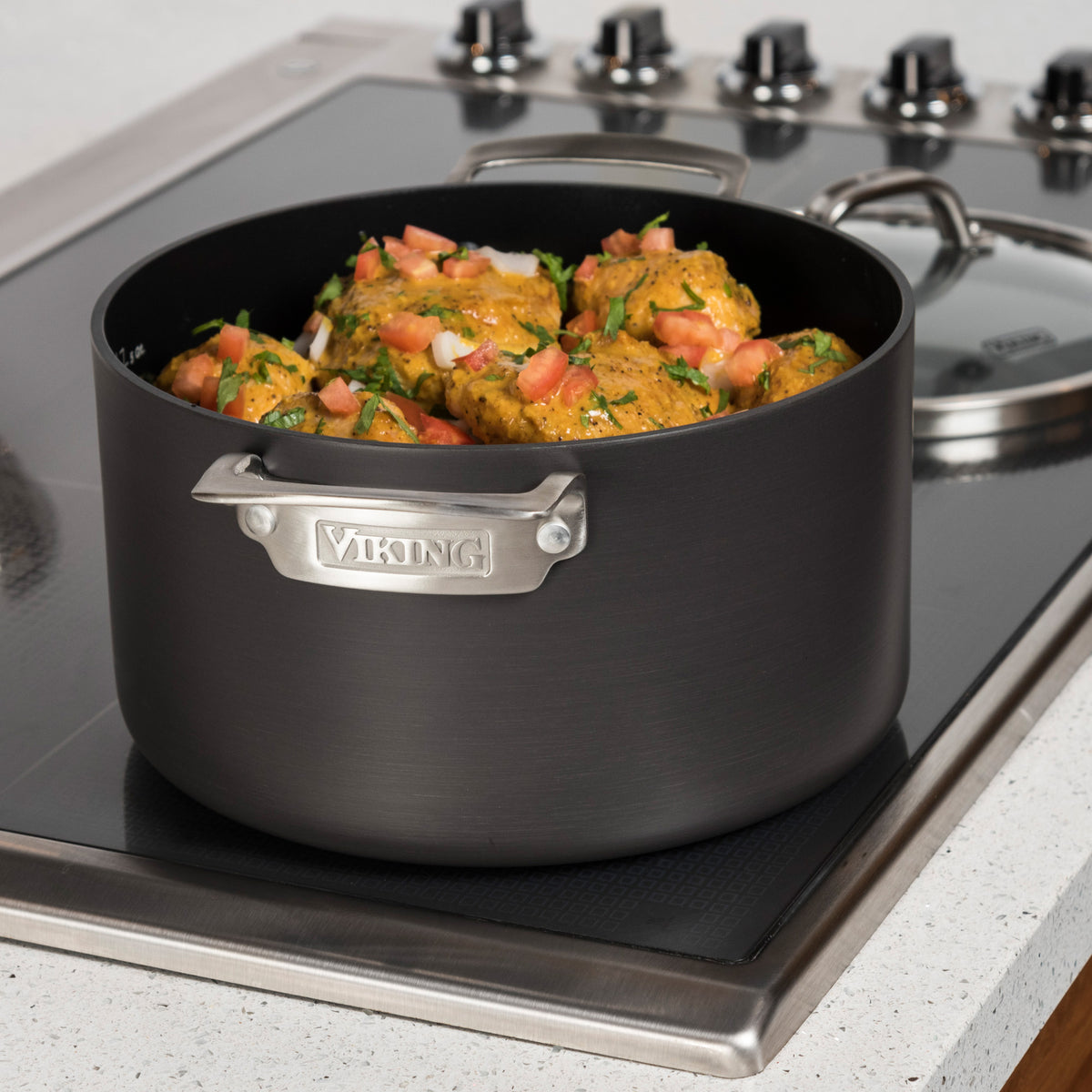 A Viking Culinary Products Hard Anodized Nonstick 6-Quart Dutch Oven with glass lid sits on a stovetop, filled with cooked chicken pieces garnished with chopped tomatoes and herbs.