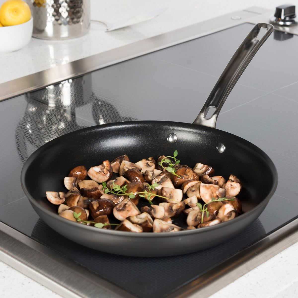 A Viking Culinary Products Hard Anodized Nonstick frying pan from the 10-piece set is shown on a stovetop in a modern kitchen, cooking chopped mushrooms and fresh herbs under glass lids.