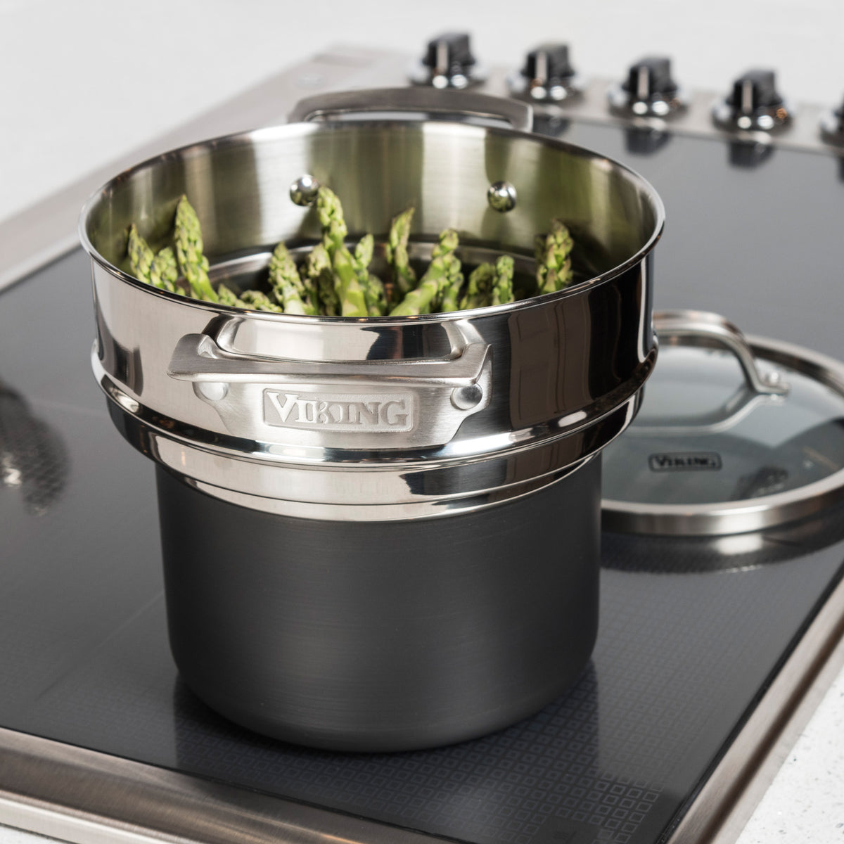 A Viking Culinary Products Stainless Steel Universal Steamer Insert and Colander holds asparagus spears on a stovetop; its glass lid is nearby. The dishwasher-safe insert is labeled Viking.
