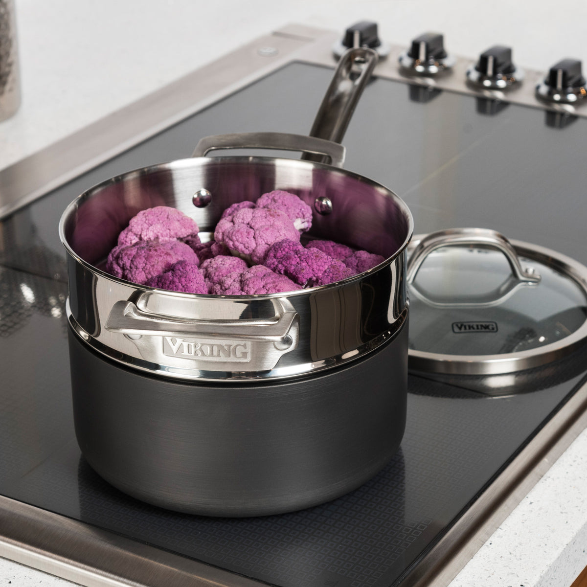 A Viking Stainless Steel Universal Steamer Insert and Colander by Viking Culinary Products holds purple cauliflower on a sleek black stovetop with multiple control knobs; the glass lid rests nearby.