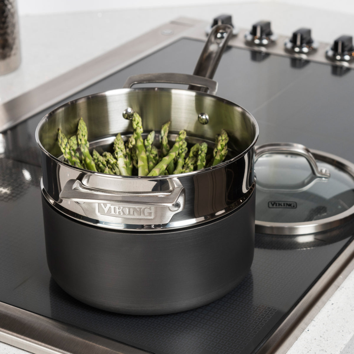 The Viking Stainless Steel Universal Steamer Insert and Colander by Viking Culinary Products rests beside a black hard anodized saucepan filled with upright asparagus on a sleek stovetop, its matching glass lid nearby.