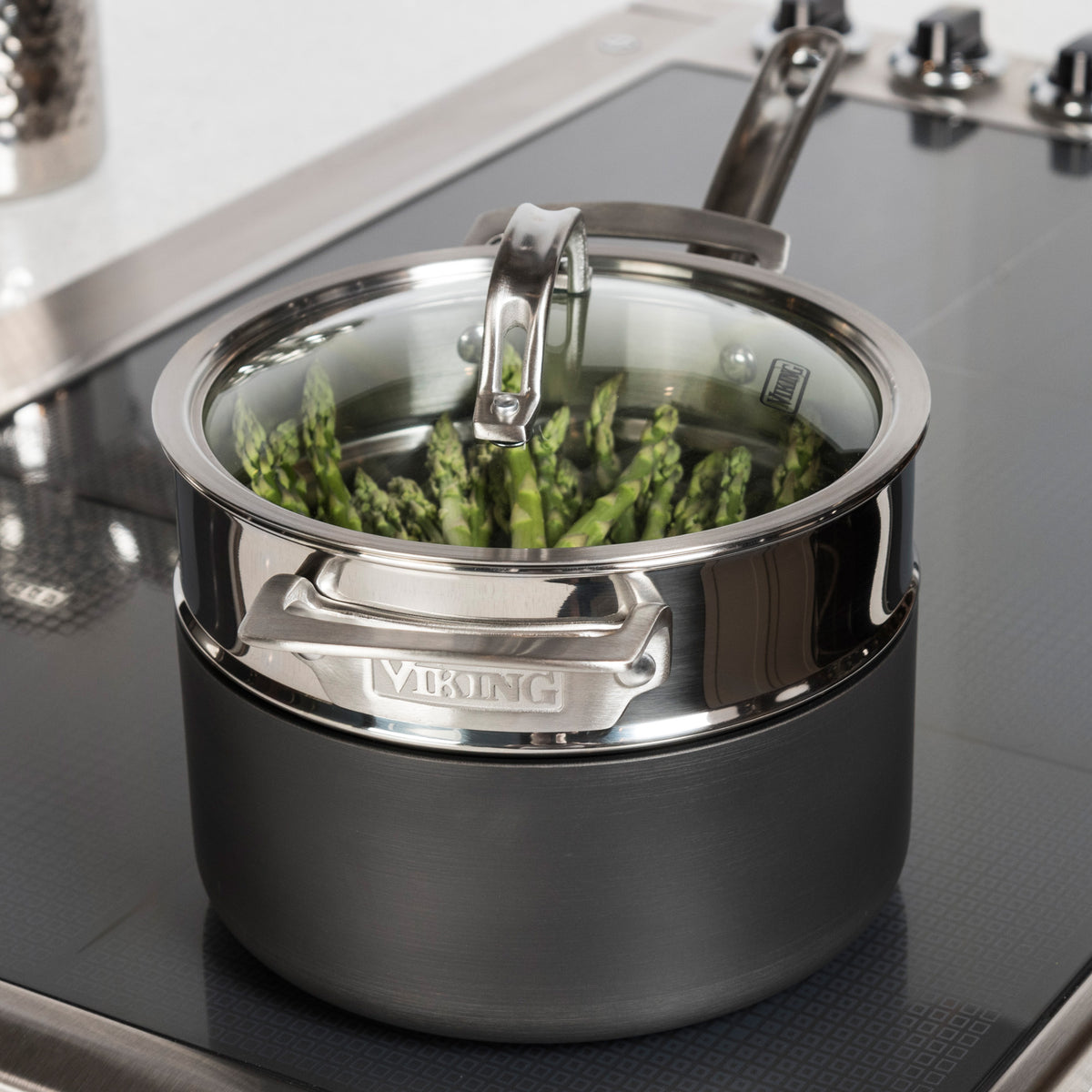 The Viking Stainless Steel Universal Steamer Insert and Colander by Viking Culinary Products steams asparagus on the stovetop, featuring a glass lid, hard anodized exterior, and dishwasher-safe design for easy cleanup.