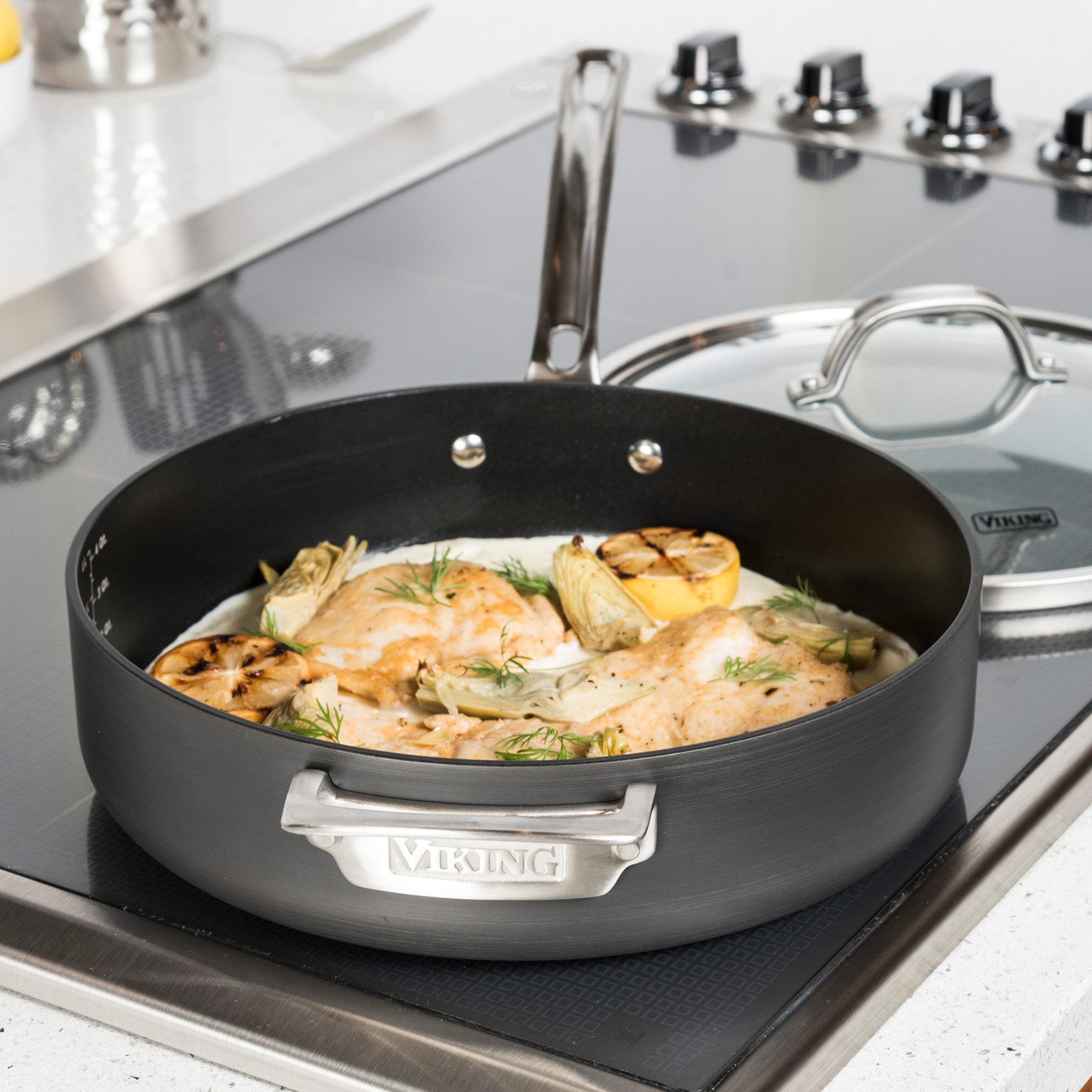 Chicken breasts, artichokes, lemon, and herbs cook in a Viking Culinary Products Hard Anodized Nonstick pan from the 10-Piece Cookware Set with Glass Lids; the glass lid rests beside the pan’s signature handles on a sleek stovetop.