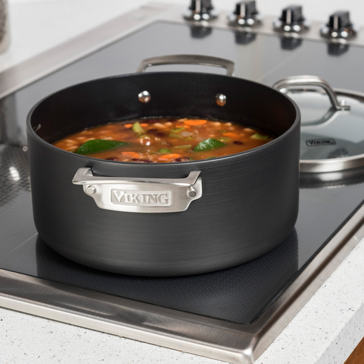 A Viking Culinary Products Hard Anodized Nonstick Dutch Oven from the 10-Piece Cookware Set sits on the stovetop, filled with soup, vegetables, and lime slices. Its sturdy handles are visible and a glass lid rests nearby.