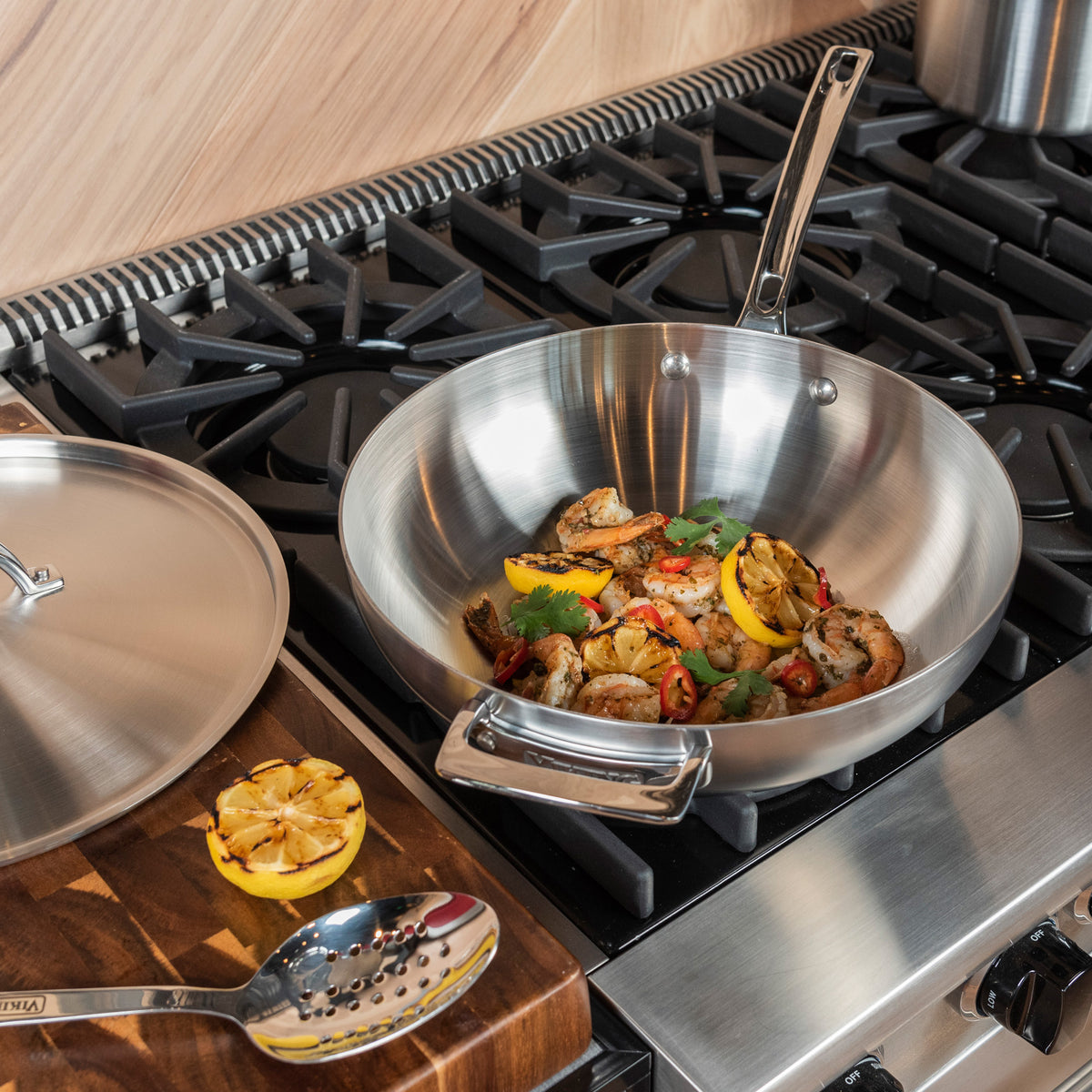 A Viking Culinary Products Professional 5-Ply 3-Quart Saucier with metal lid sits on a gas stove, filled with cooked shrimp, sliced peppers, grilled lemon, and herbs. Nearby on a wooden cutting board are a slotted spoon and grilled lemon halves.