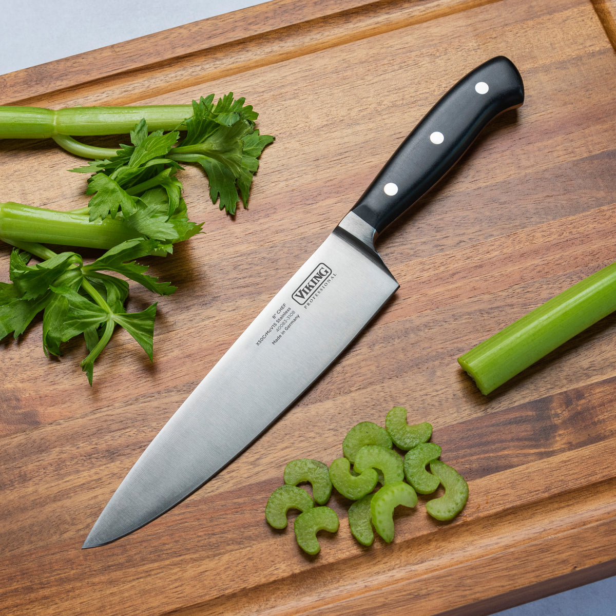 The Viking Professional 8-Inch Chefs Knife by Viking Culinary Products, featuring a black handle and durable German steel, lies on a wooden cutting board amid chopped celery and leafy stalks.