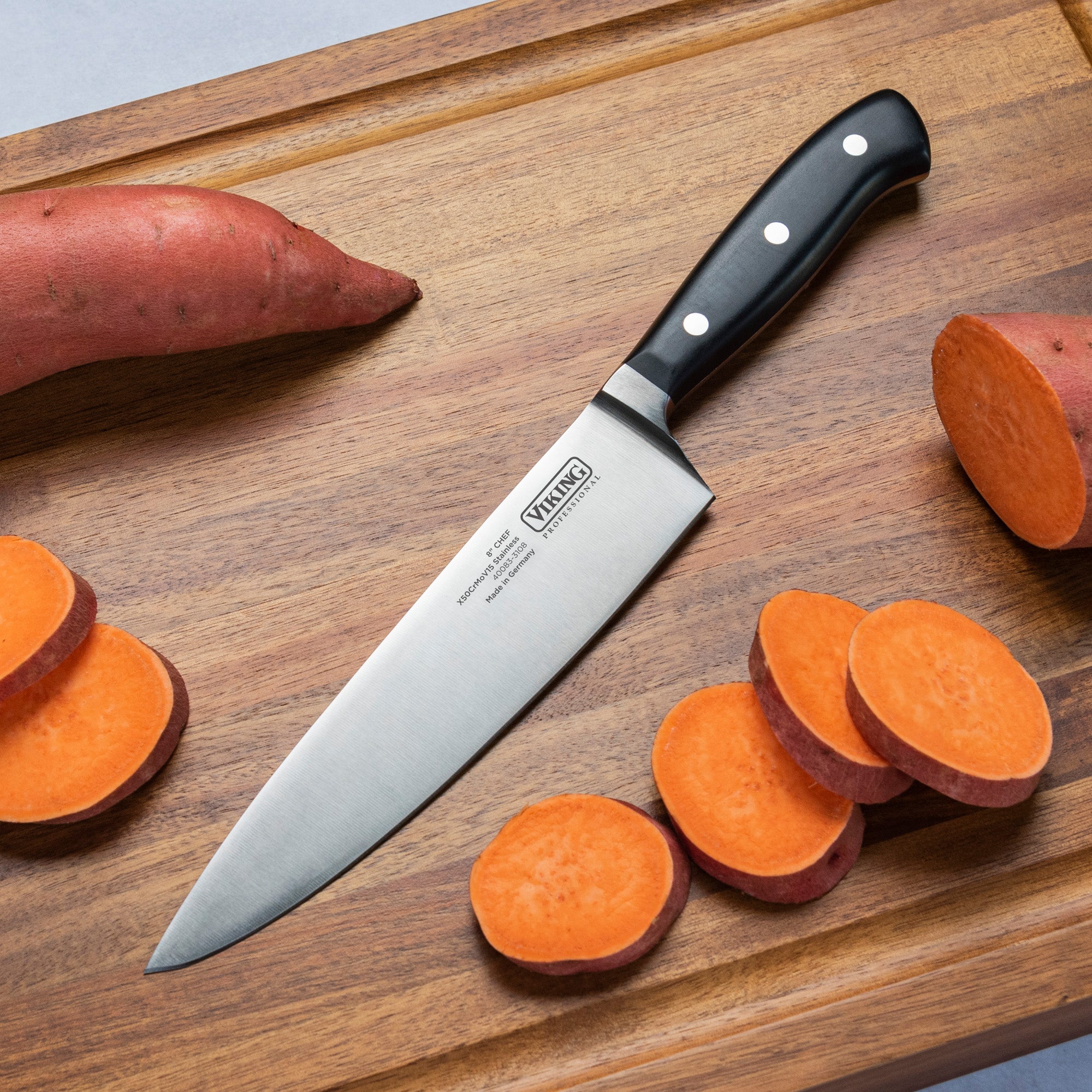 The Viking Professional 8-Inch Chef’s Knife by Viking Culinary Products, featuring a black handle and German steel blade, rests on a wooden cutting board with whole and sliced sweet potatoes ready for meal prep.
