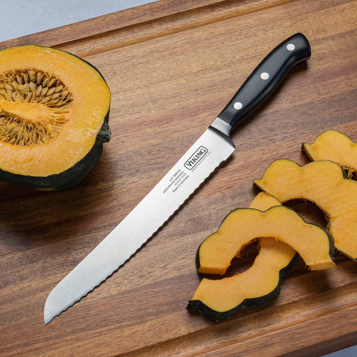 The Viking Professional 8-Inch Bread Knife by Viking Culinary Products, featuring a black handle and full-tang serrated blade, sits on a wooden cutting board beside a halved squash and several squash slices.
