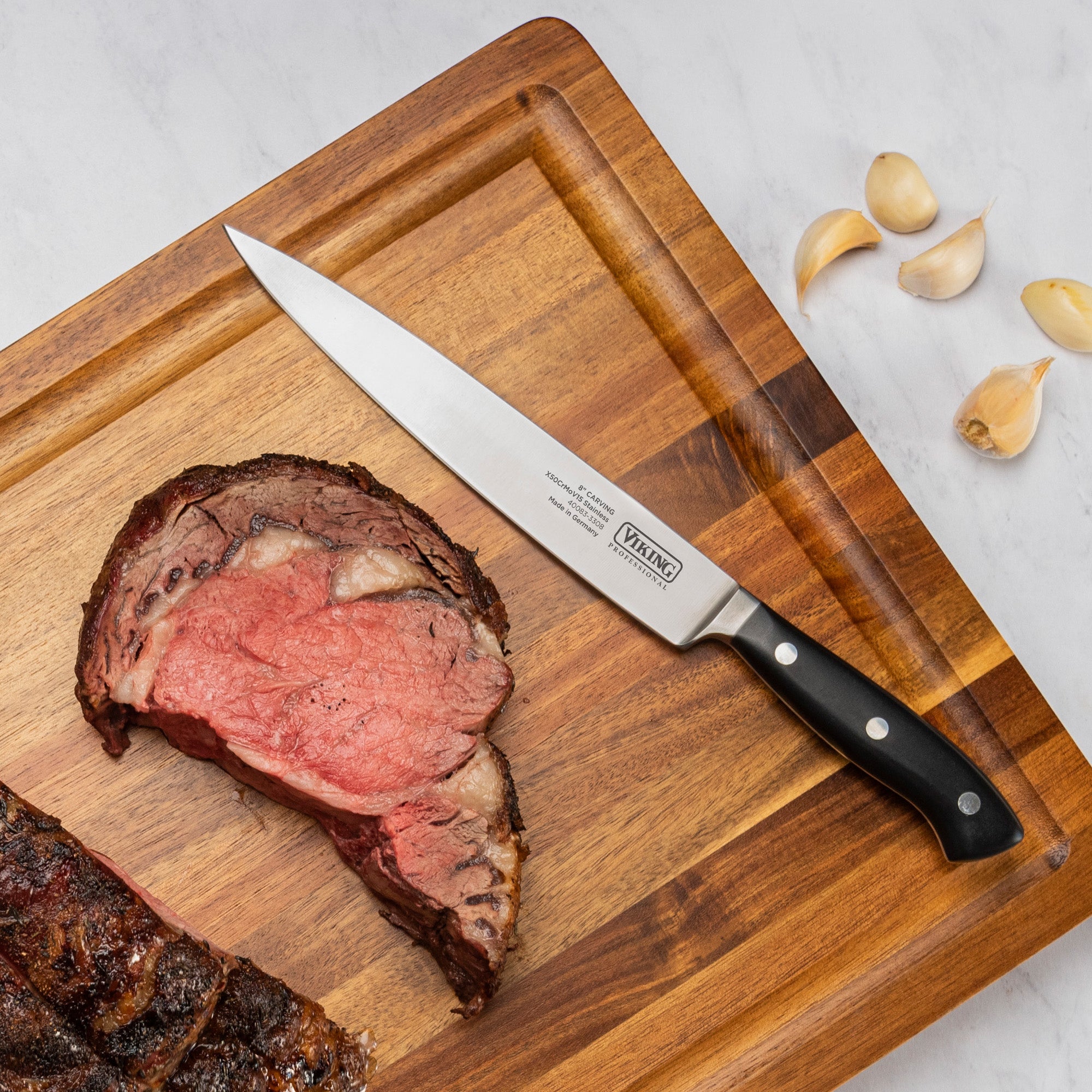 A Viking Professional 8 Carving Knife by Viking Culinary Products lies on a wooden cutting board beside a thick slice of medium-rare roast beef, with several garlic cloves nearby on a white marble surface.