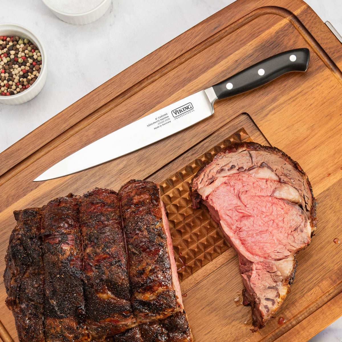 A juicy roast beef sits on a wooden board beside the Viking Professional 2-Piece Carving Set by Viking Culinary Products. A thick slice reveals the pink center, with a small bowl of peppercorns nearby.