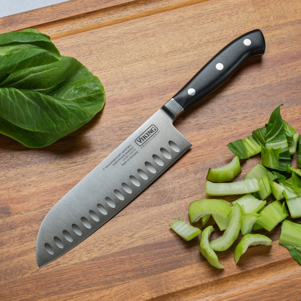 The Viking Professional 7-Inch Santoku Knife from Viking Culinary Products, featuring a black handle and crafted from high-quality German steel, rests on a wooden cutting board beside leafy greens and chopped celery.