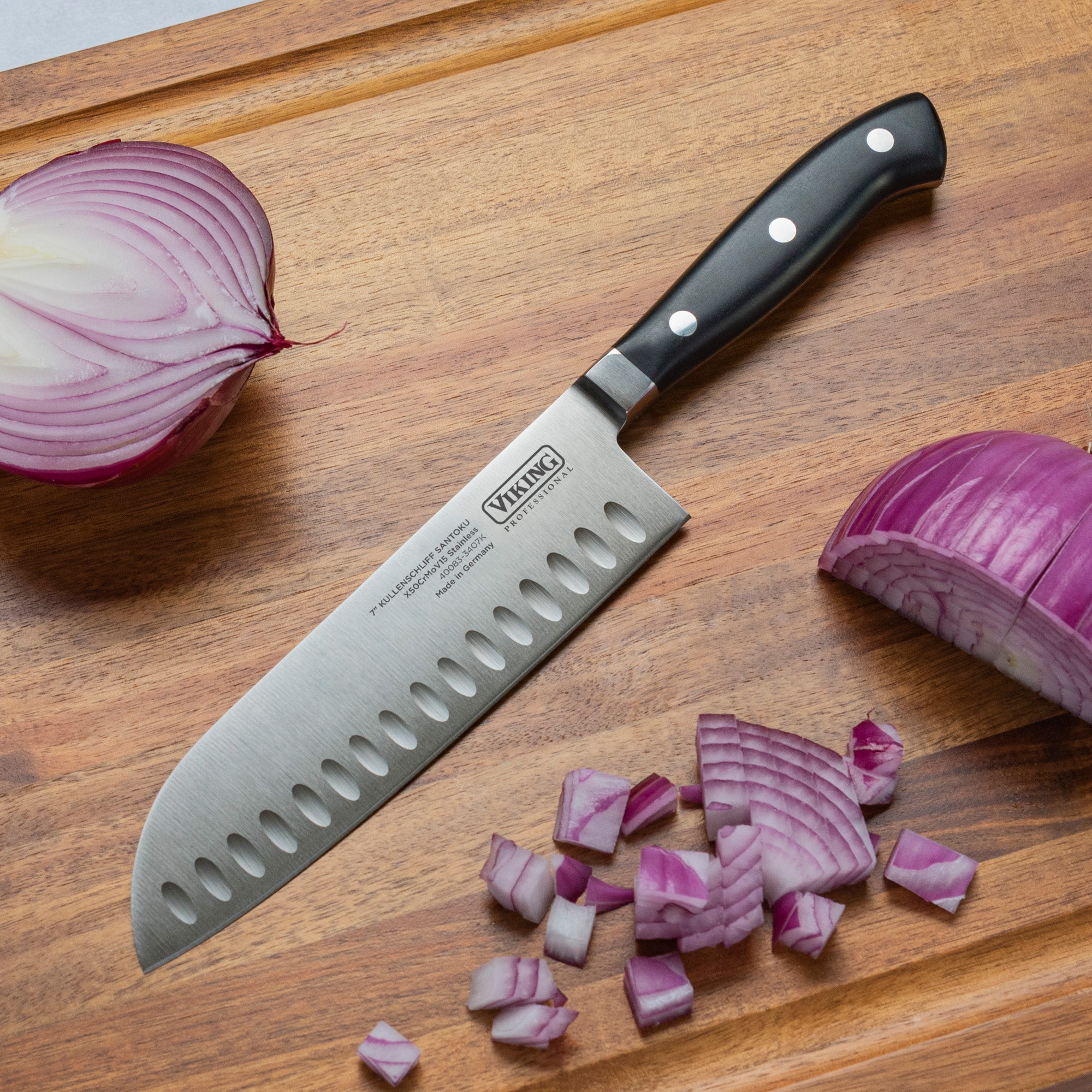 A Viking Professional 7-Inch Santoku Knife by Viking Culinary Products rests on a wooden cutting board beside a halved red onion, a quartered red onion, and several diced pieces of red onion.