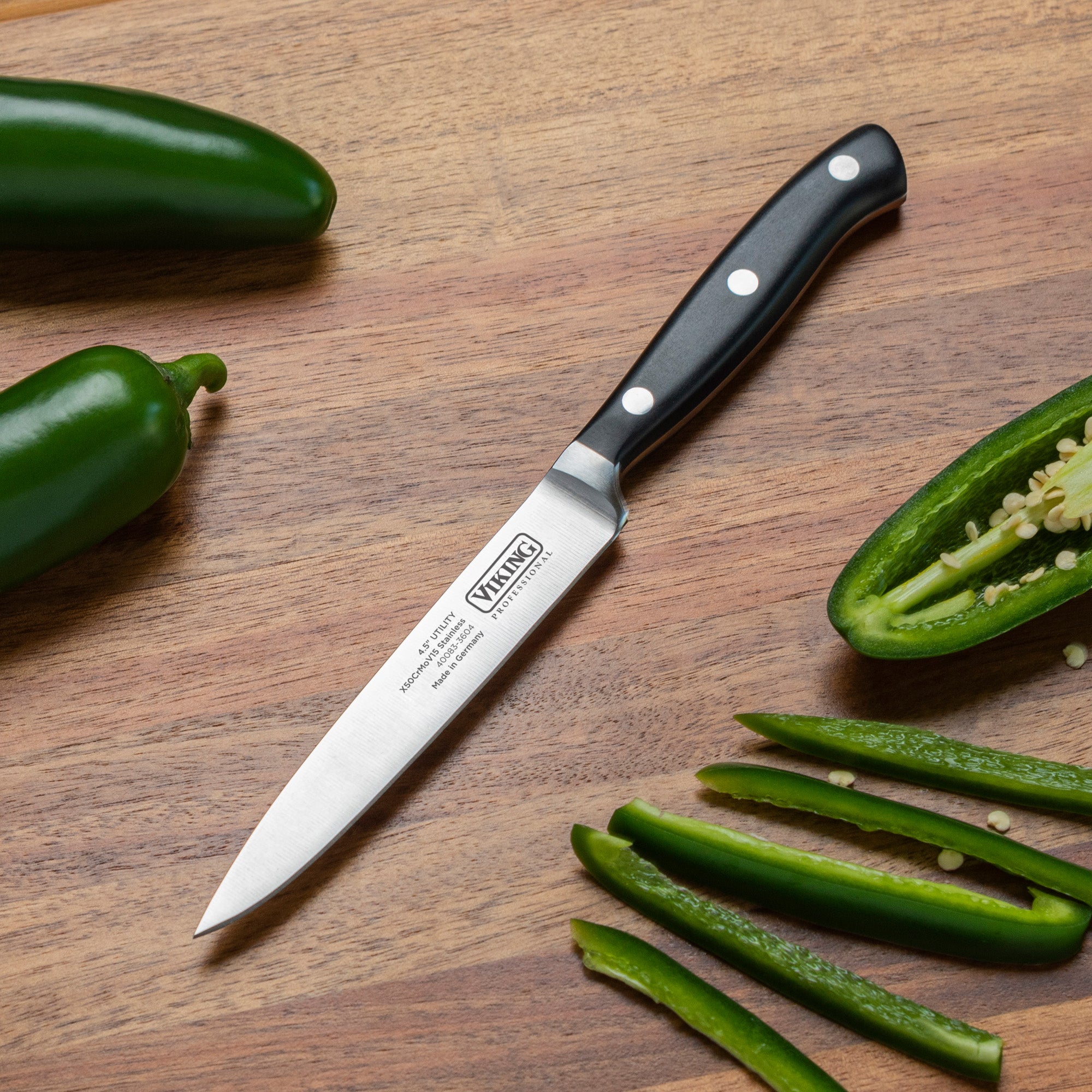 A Viking Professional 4.5-Inch Utility Knife by Viking Culinary Products, featuring a black handle and German steel blade, rests on a wooden surface with whole and sliced jalapeños, seeds visible and slices neatly arranged.