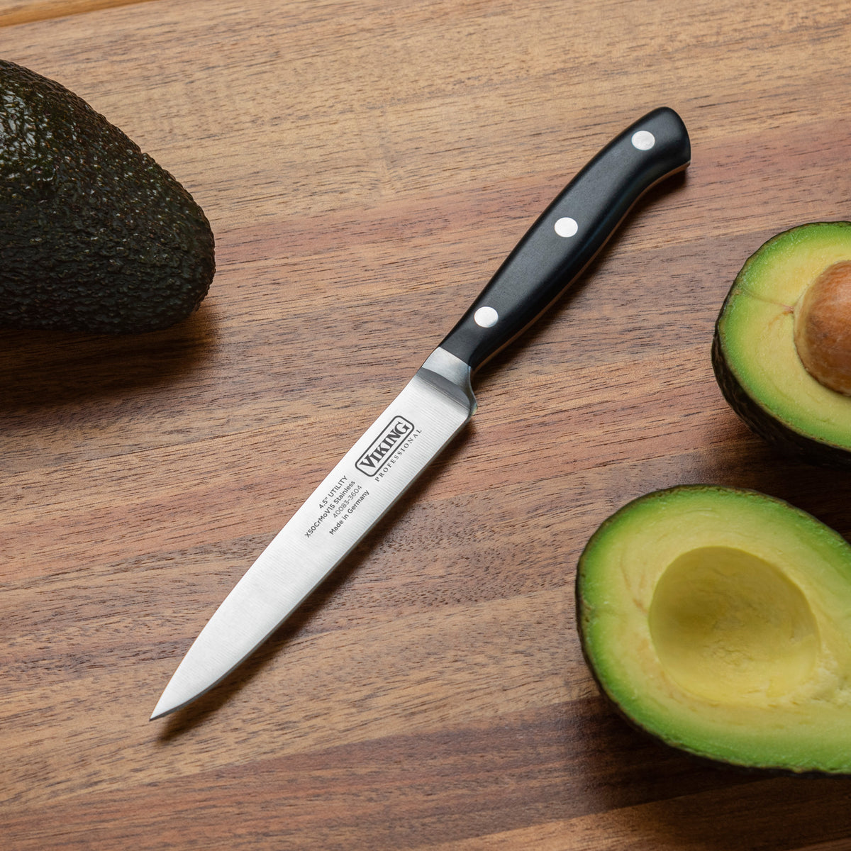The Viking Professional 4.5-Inch Utility Knife by Viking Culinary Products, featuring a black handle and German steel blade, rests on a wooden surface next to a whole avocado and a halved avocado with its pit visible.