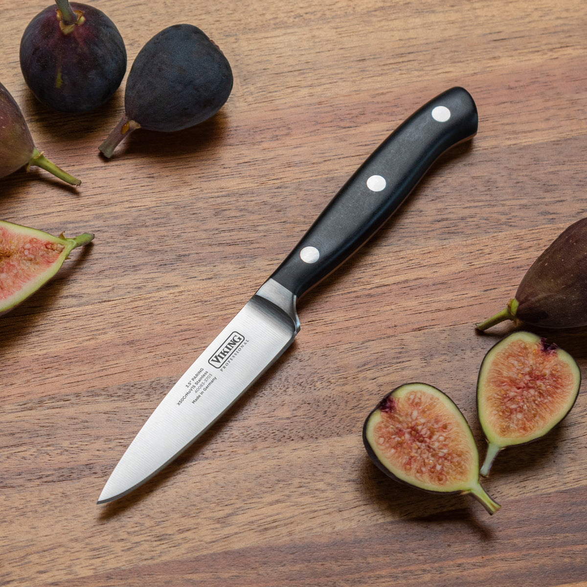 A Viking Professional 3.5-Inch Paring Knife by Viking Culinary Products with a black triple-riveted handle rests on a wooden surface amid whole and sliced figs.
