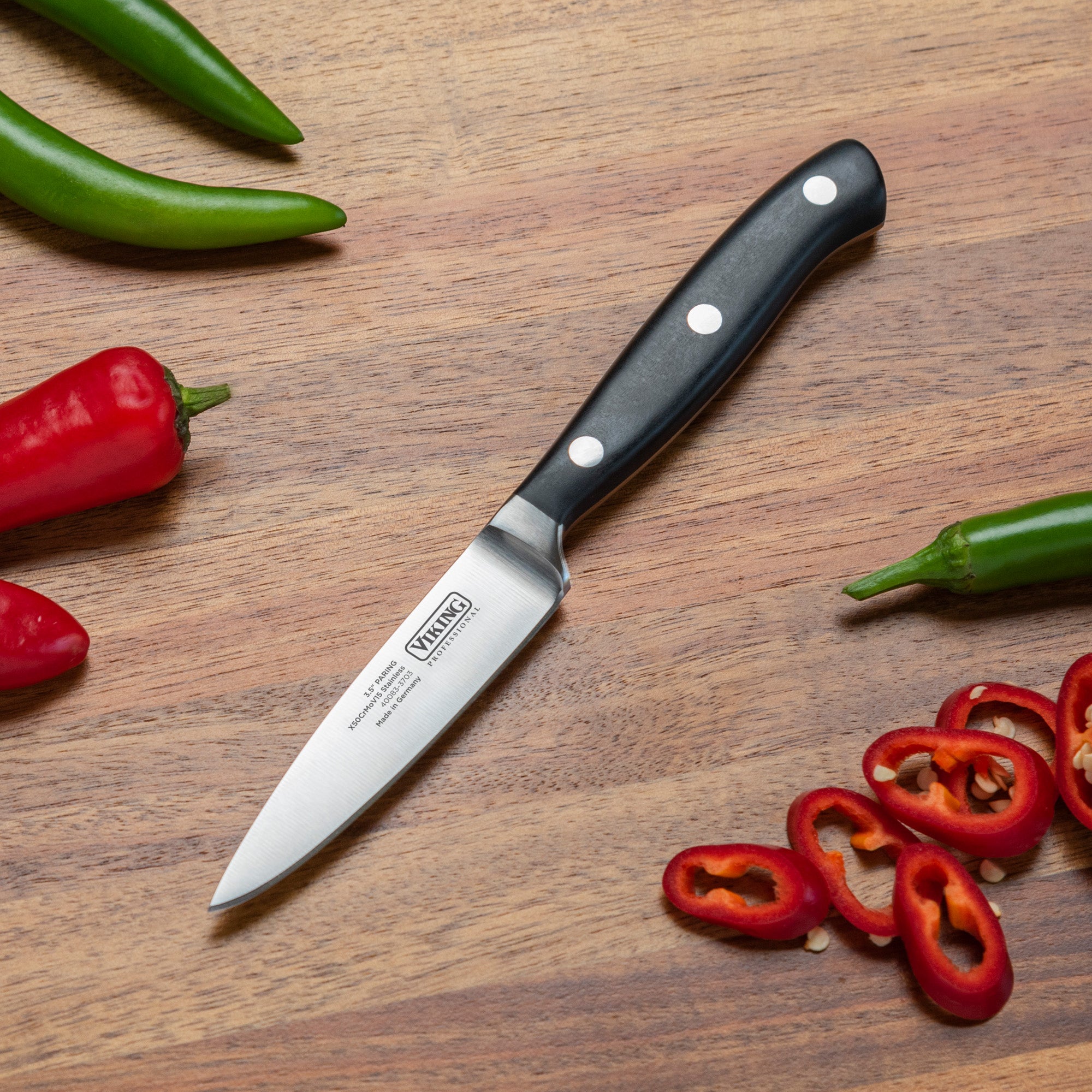 A Viking Professional 3.5-Inch Paring Knife by Viking Culinary Products with a black handle rests on wood, surrounded by whole green and red chili peppers and sliced red chili pieces.