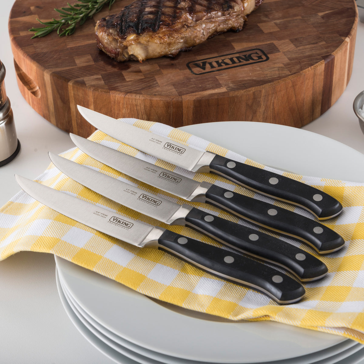 The Viking Professional 4-Piece Steak Knife Set by Viking Culinary Products, featuring black handles and full-tang design, is displayed on a yellow and white checkered cloth over stacked white plates with a steak and cutting board in the background.