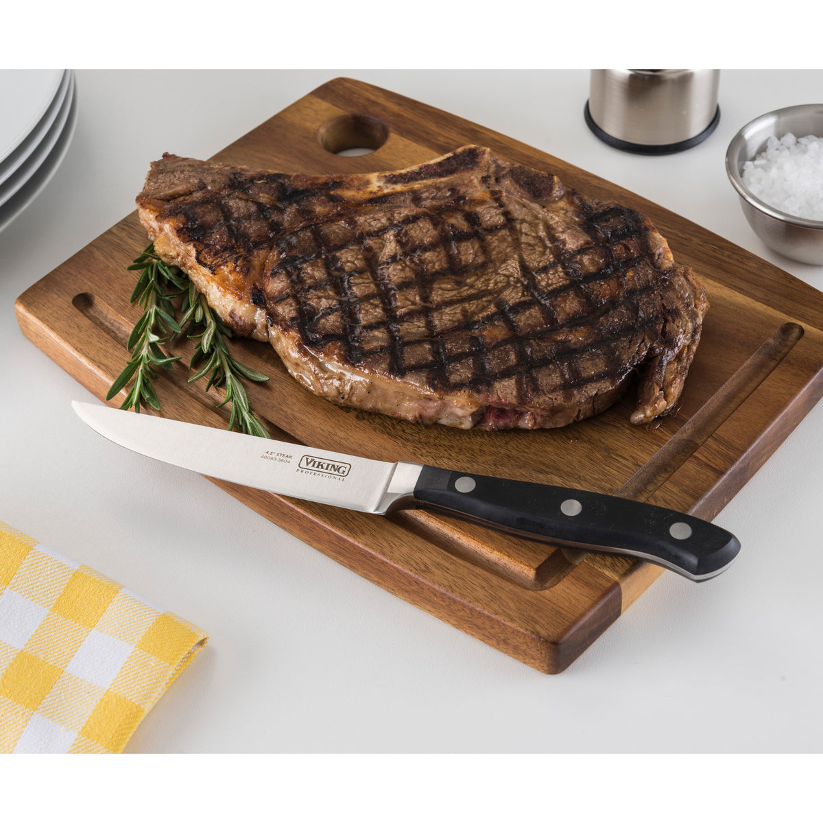 A grilled steak with char marks rests on a wooden cutting board beside the Viking Professional 4.5-Inch Steak Knife by Viking Culinary Products, garnished with rosemary. Plates, a yellow check napkin, coarse salt bowl, and pepper grinder are nearby.