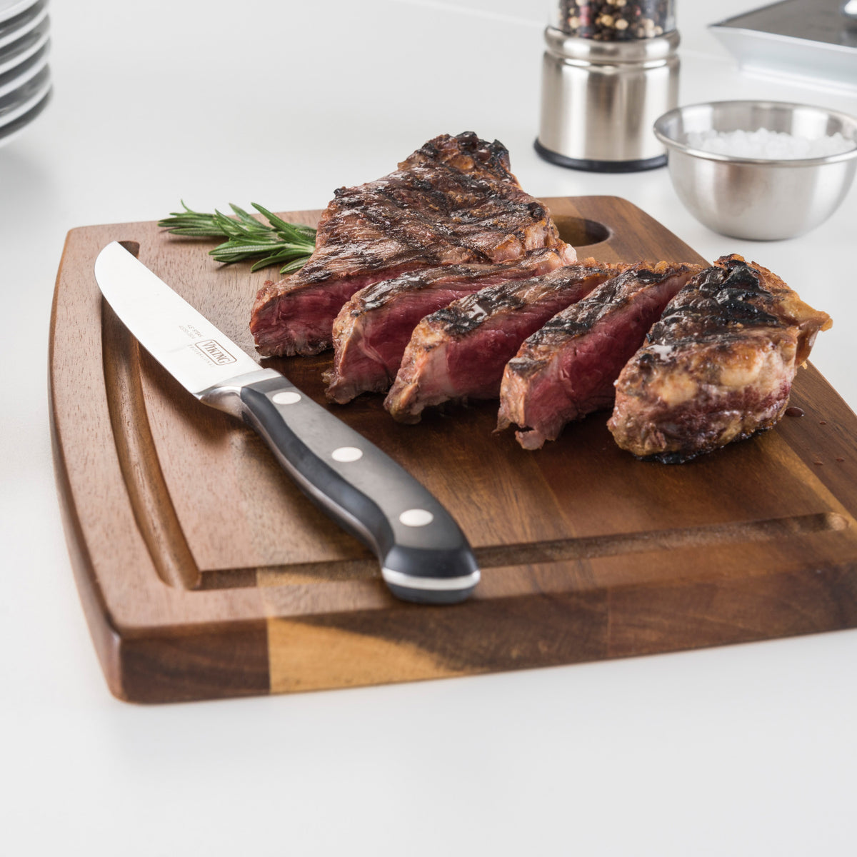 A grilled steak, sliced to rare and set on a wooden board, is accompanied by a Viking Professional Steak Knife from Viking Culinary Products with full-tang design and rosemary. A metal bowl of salt and pepper grinder appear in the background.