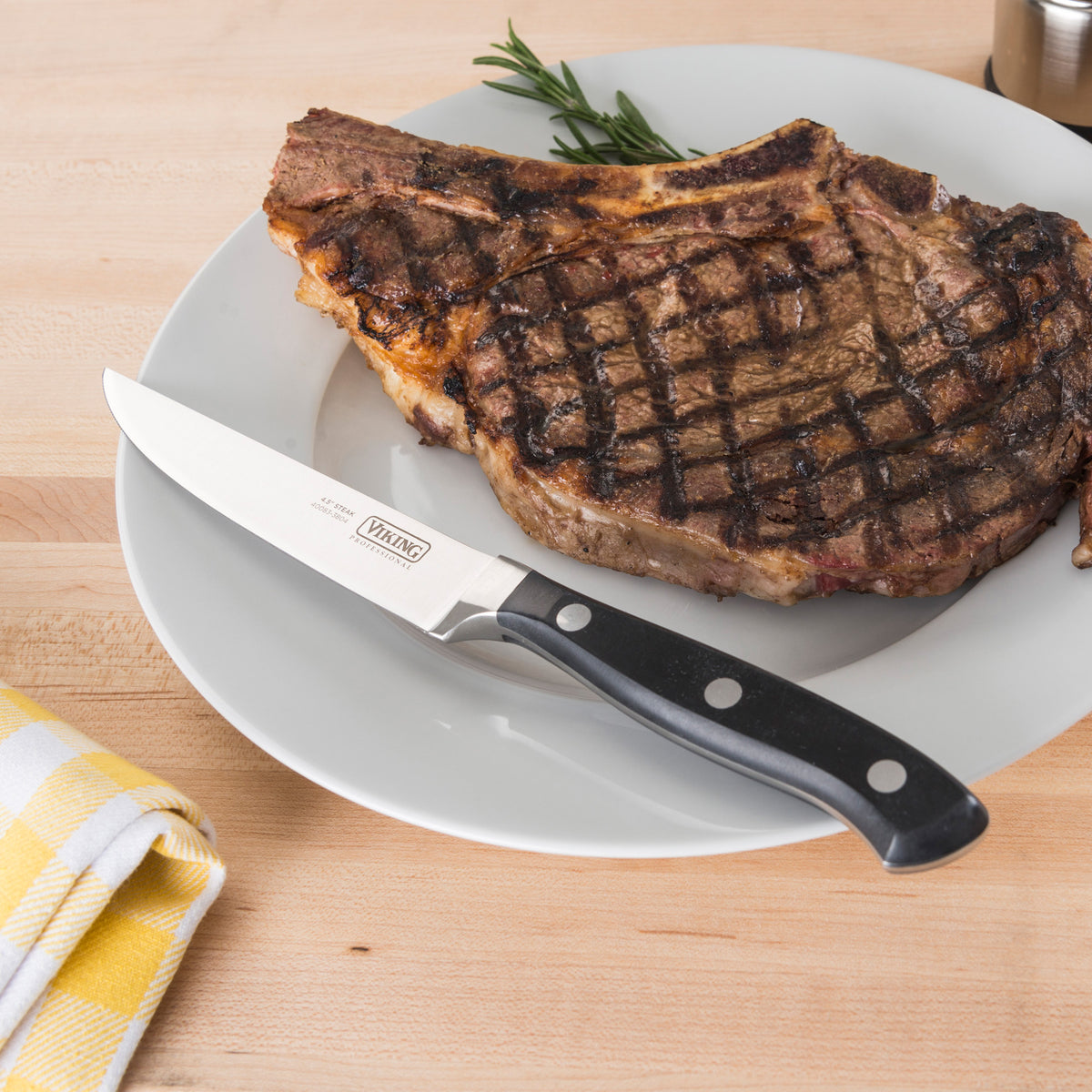 A grilled T-bone steak with sear marks sits on a white plate next to a Viking Professional 4.5-Inch Steak Knife by Viking Culinary Products, garnished with rosemary and a yellow checkered napkin on a wooden surface.