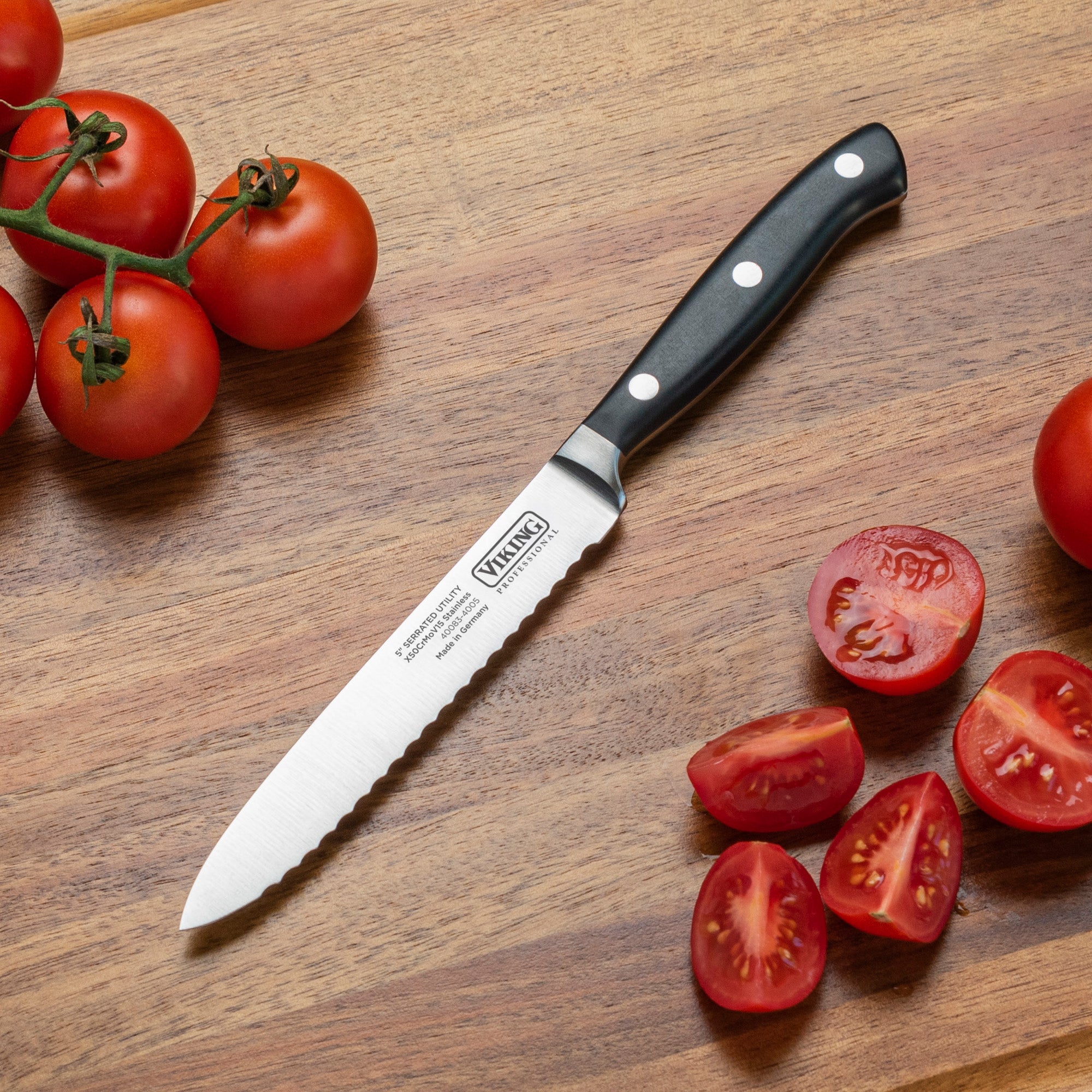 A Viking Professional 5-Inch Serrated Utility Knife by Viking Culinary Products, with a black handle, lies on a wooden cutting board surrounded by whole vine tomatoes and sliced tomatoes.