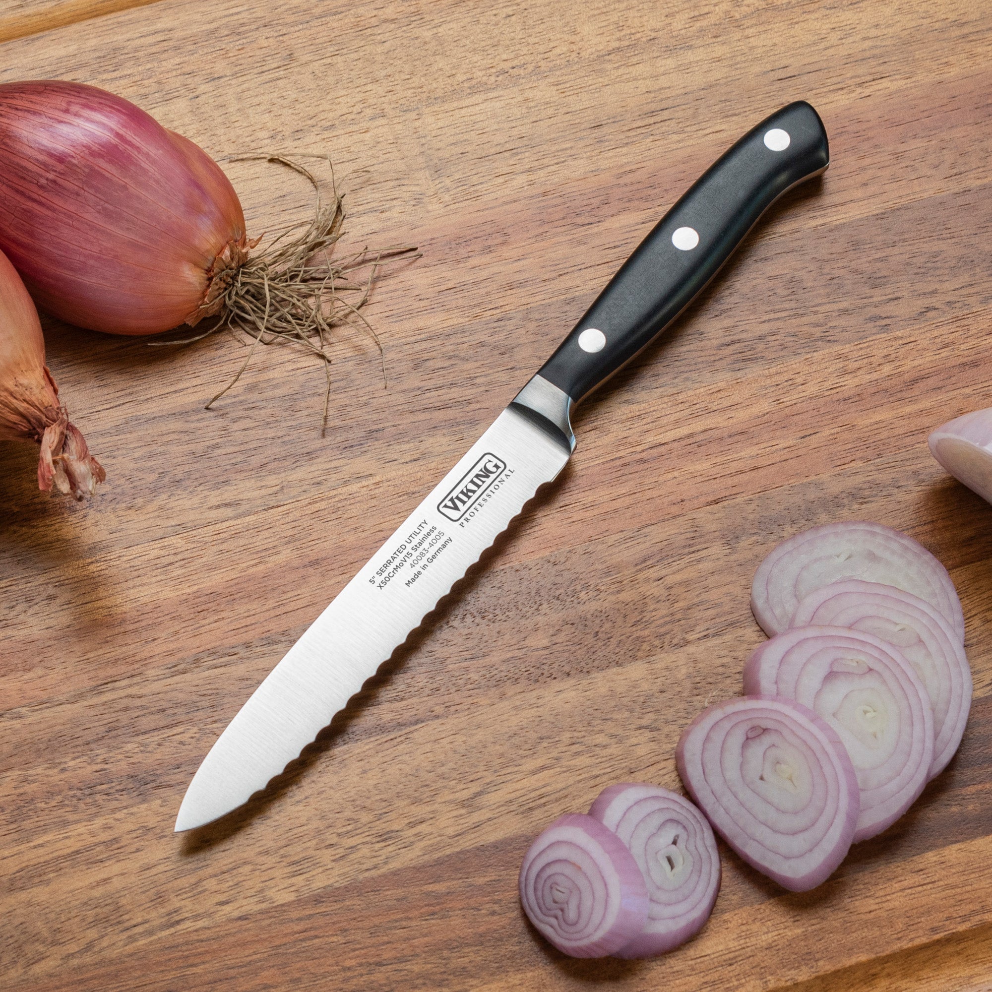 A Viking Professional 5-Inch Serrated Utility Knife by Viking Culinary Products with a black handle rests on a wooden cutting board beside sliced and whole shallots.