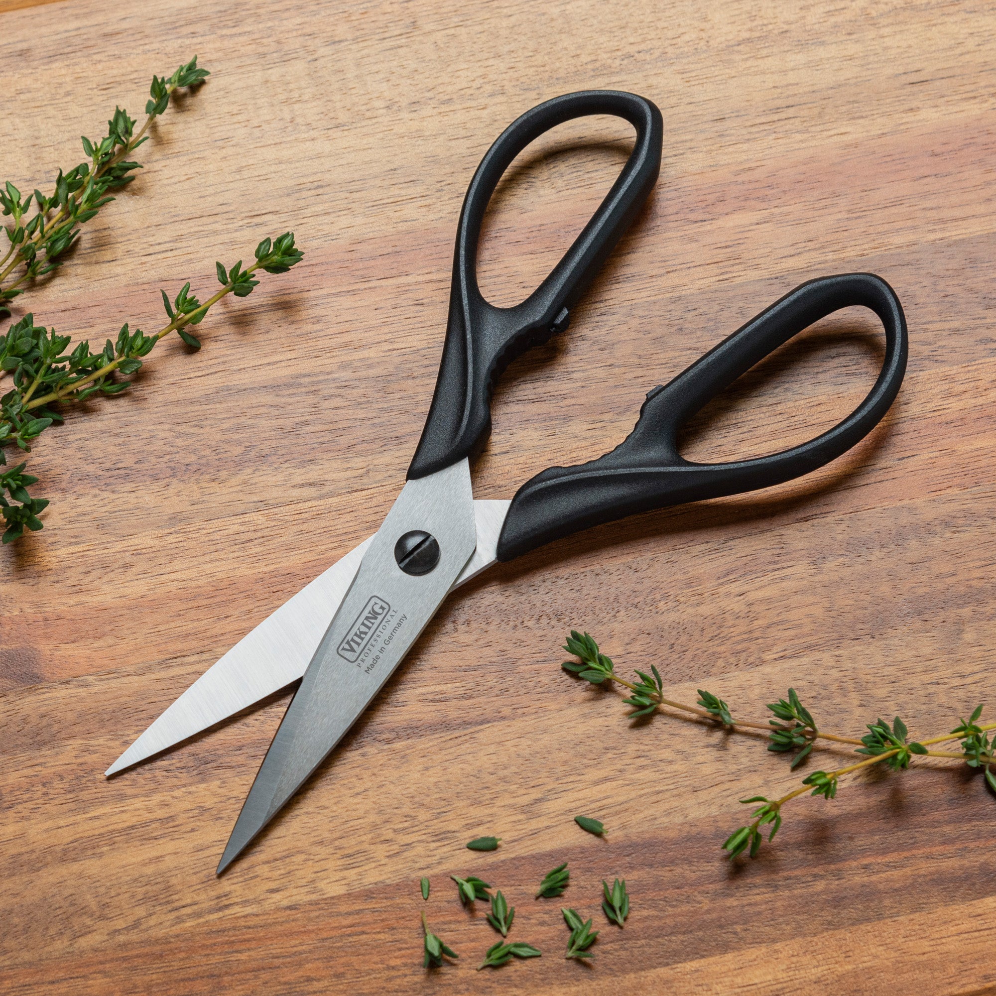 A pair of Viking Professional 8-Inch Scissors by Viking Culinary Products, featuring black handles and a full-tang design, rests on a wooden surface surrounded by small sprigs of fresh green herbs.