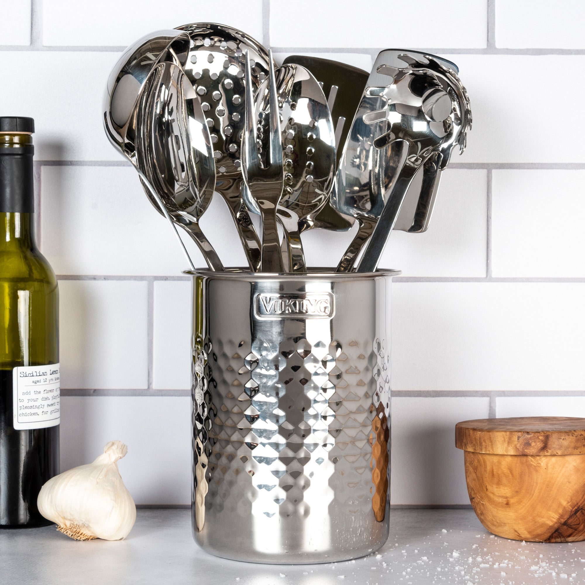 The Viking Hammered Stainless Steel Utensil Holder by Viking Culinary Products, filled with metal kitchen utensils, sits on the counter beside olive oil, garlic, and a wooden container against a white tiled wall.