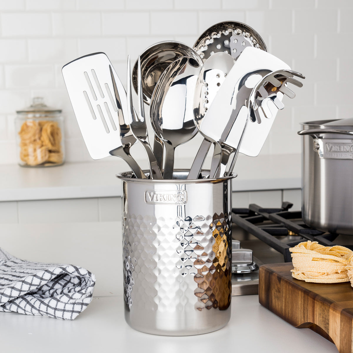 The Viking Hammered Stainless Steel Utensil Holder by Viking Culinary Products sits on the countertop near the stove, filled with spatulas, ladles, and serving forks. Nearby are a towel, pasta jar, and a cutting board with pasta ready to cook.