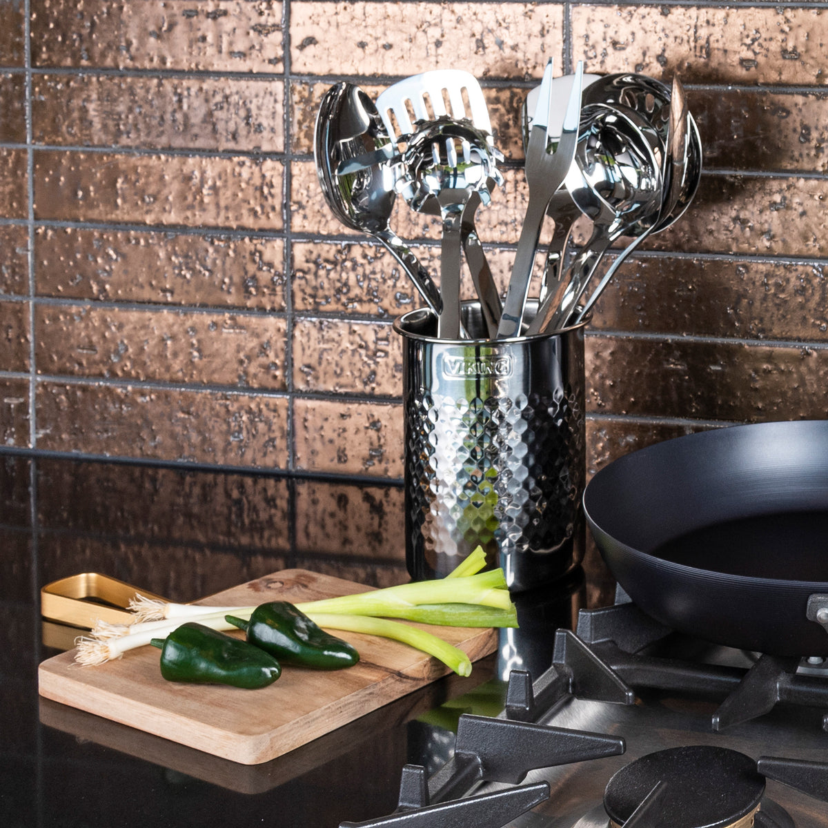 A Viking Hammered Stainless Steel Utensil Holder by Viking Culinary Products sits on a counter with kitchen tools beside a cutting board with green onions and poblano peppers, near a black skillet on the stove against brown tiled backsplash.
