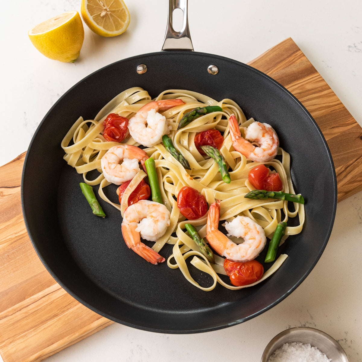 A Viking Contemporary 3-Ply Stainless Steel 12-Inch Nonstick Fry Pan by Viking Culinary Products holds fettuccine, shrimp, cherry tomatoes, and asparagus on a wooden board with a halved lemon and bowl of salt beside it on a white surface.