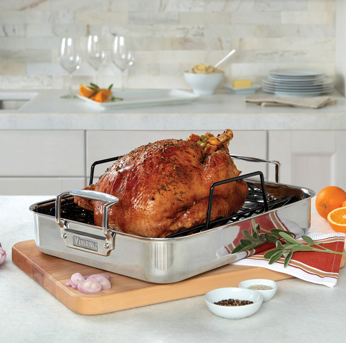 A roasted turkey garnished with herbs rests on the Viking 3-Ply Stainless Steel Roaster with Rack by Viking Culinary Products atop a wooden board, surrounded by orange slices, shallots, herbs, salt, and pepper in a modern kitchen.