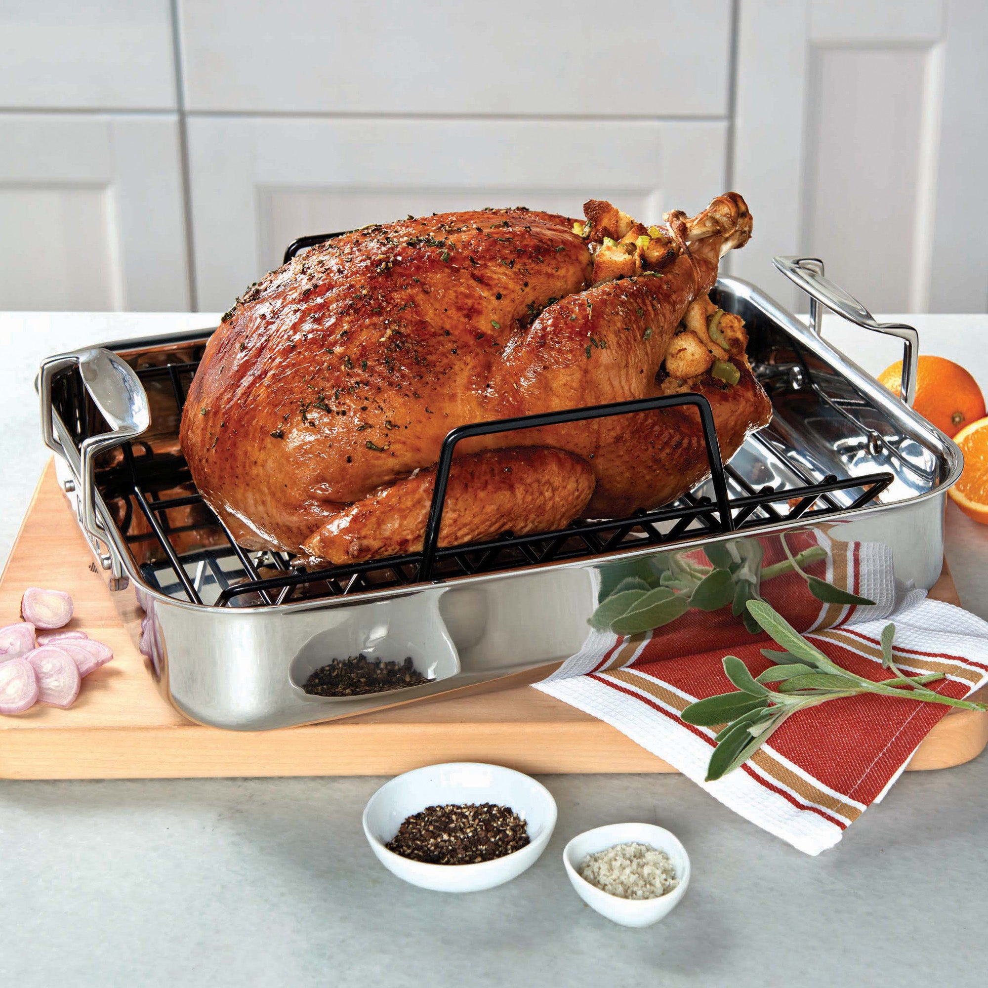 A roasted turkey rests on the Viking 3-Ply Stainless Steel Roaster with Rack by Viking Culinary Products, set on a wooden board and surrounded by fresh herbs, orange slices, shallots, and small bowls of salt and pepper.
