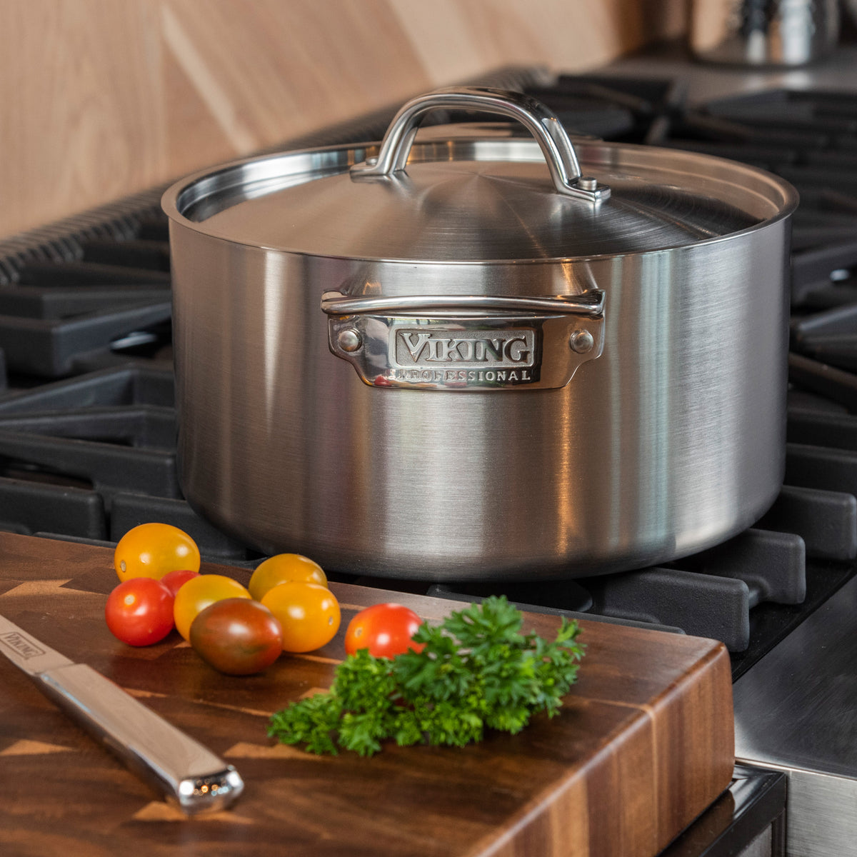 A Viking Culinary Products Professional 5-Ply Stainless Steel 6-Quart Stock Pot with stay-cool handles sits on the stove, while cherry tomatoes, a knife, and fresh parsley are arranged on a wooden cutting board in front.