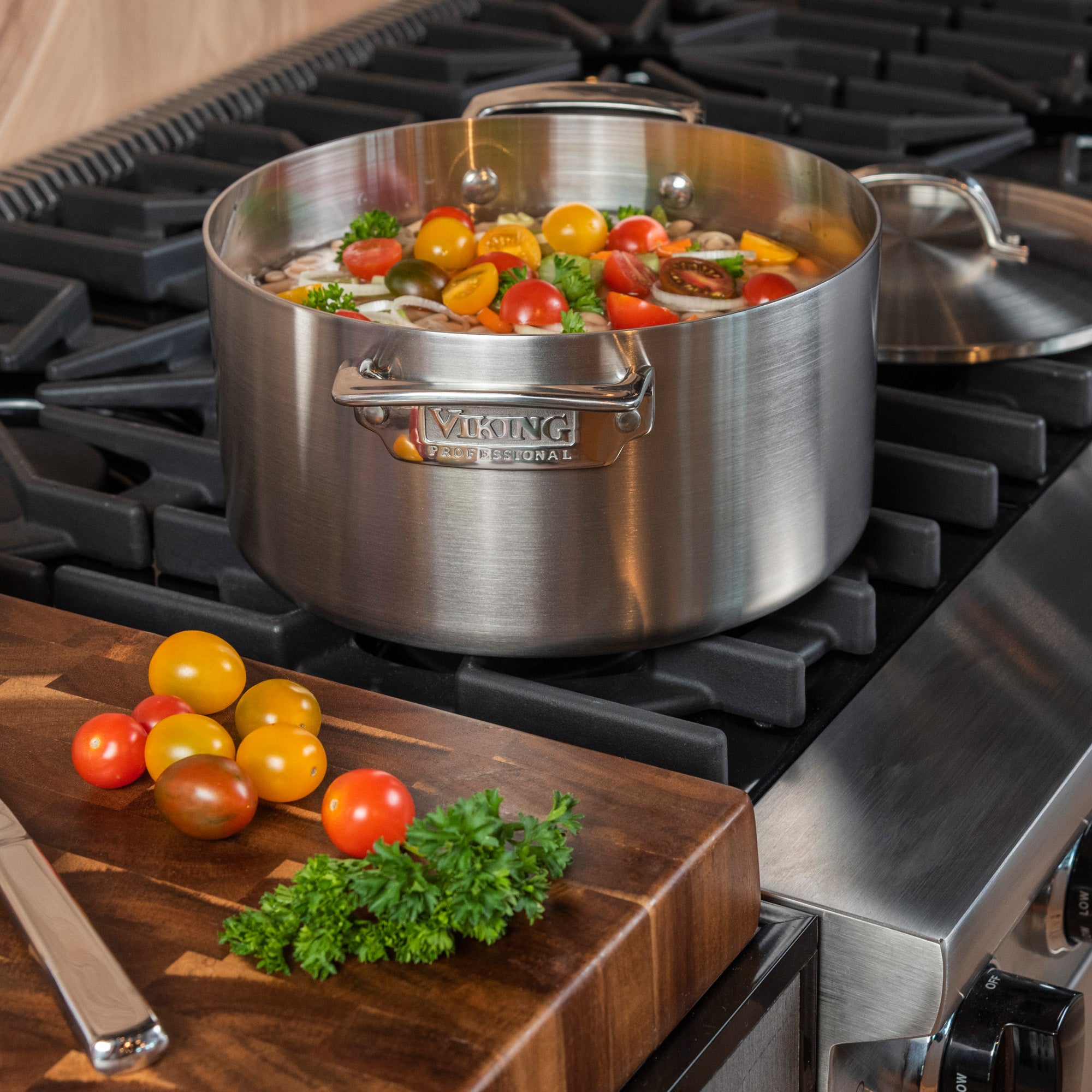 The Viking Professional 5-Ply 5-Piece Starter Cookware Set from Viking Culinary Products is shown on a gas stovetop with cherry tomatoes and vegetables inside, while a wooden cutting board with fresh tomatoes and parsley sits nearby.