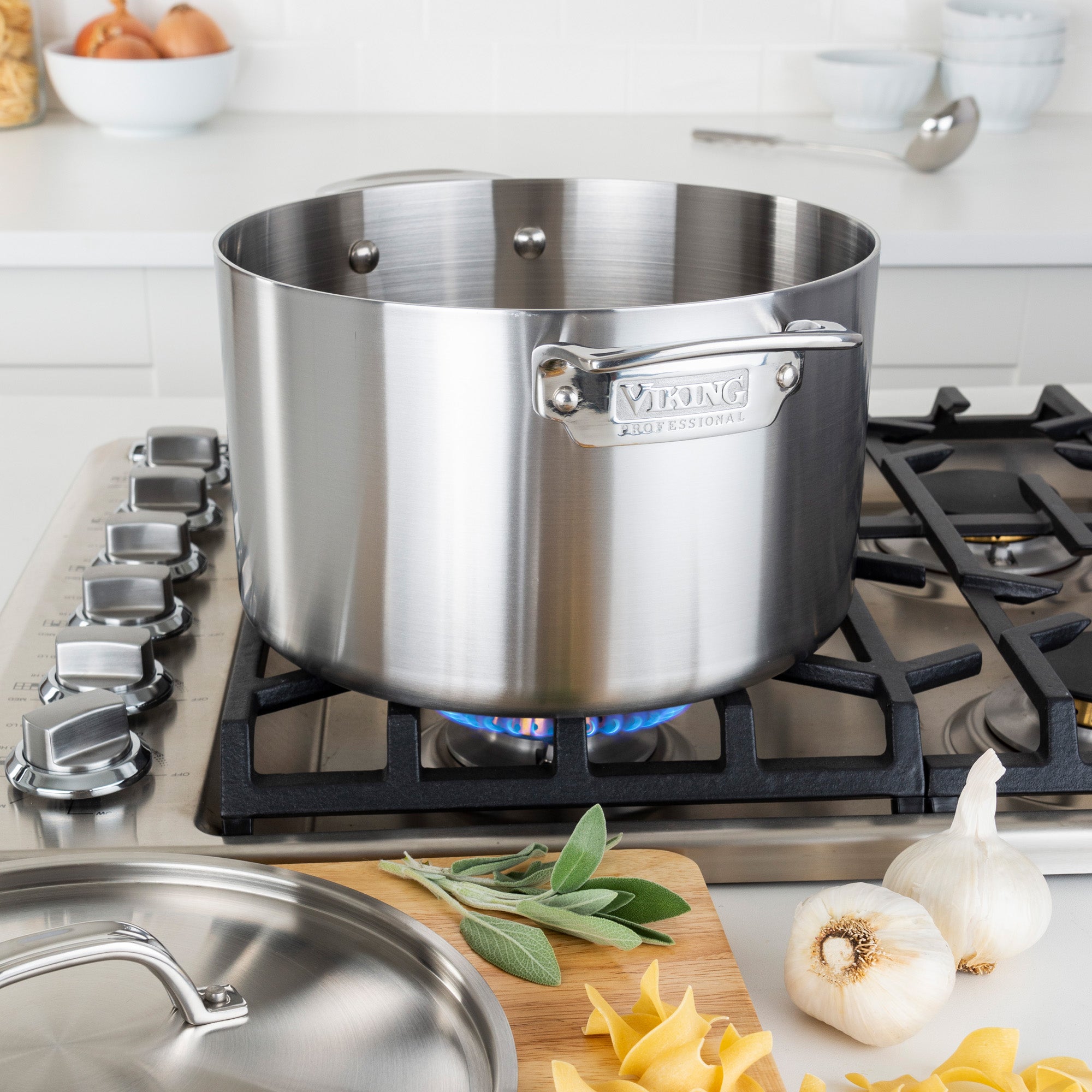 The Viking Professional 5-Ply Stainless Steel 8-Quart Stock Pot by Viking Culinary Products sits on a lit gas stove in a modern kitchen, surrounded by fresh herbs, garlic, and uncooked pasta, with its lid resting on the counter.