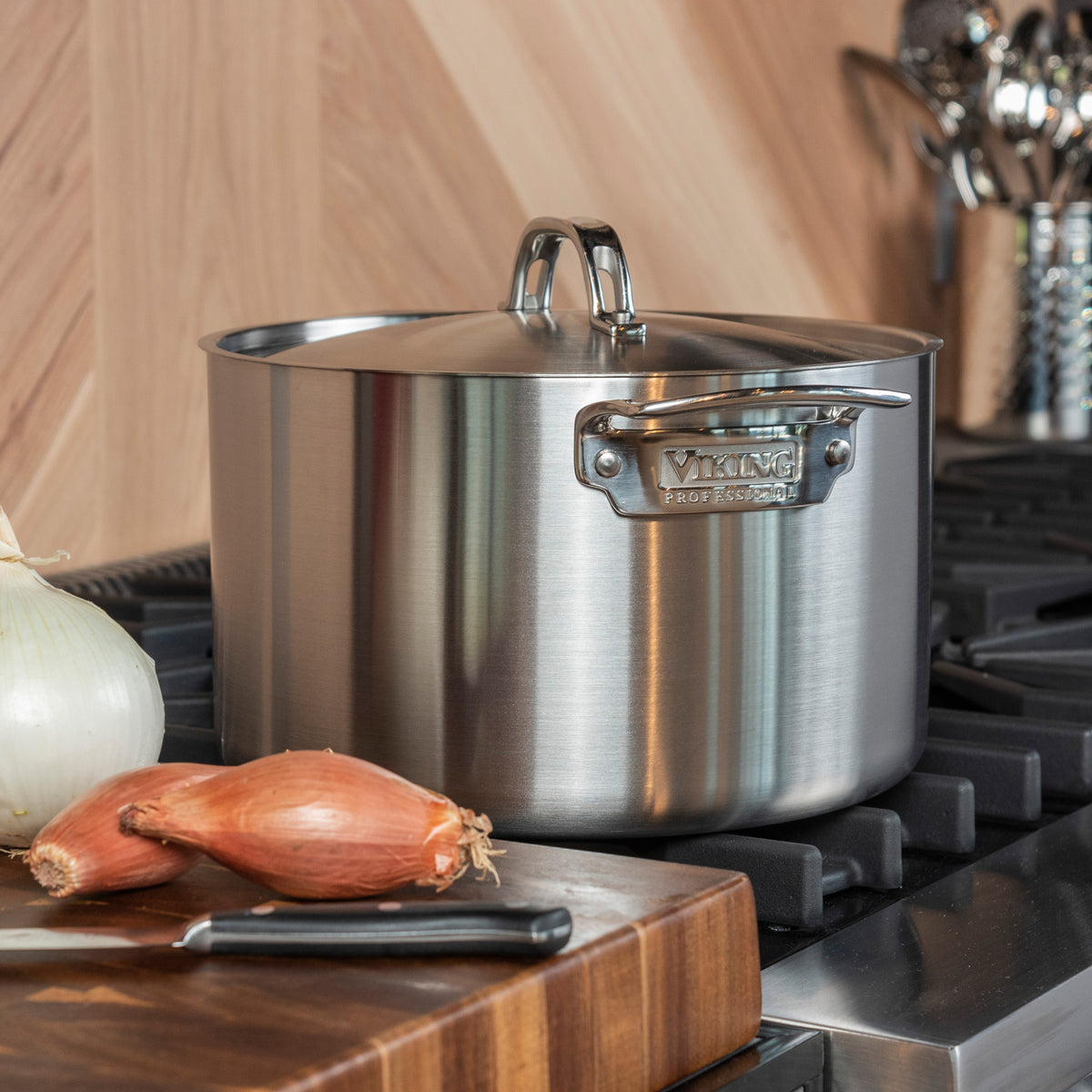 A Viking Professional 5-Ply Stainless Steel 8-Quart Stock Pot by Viking Culinary Products with lid sits on a stove; in front, a cutting board displays a knife, peeled shallot, and onion. Kitchen utensils appear in the background.
