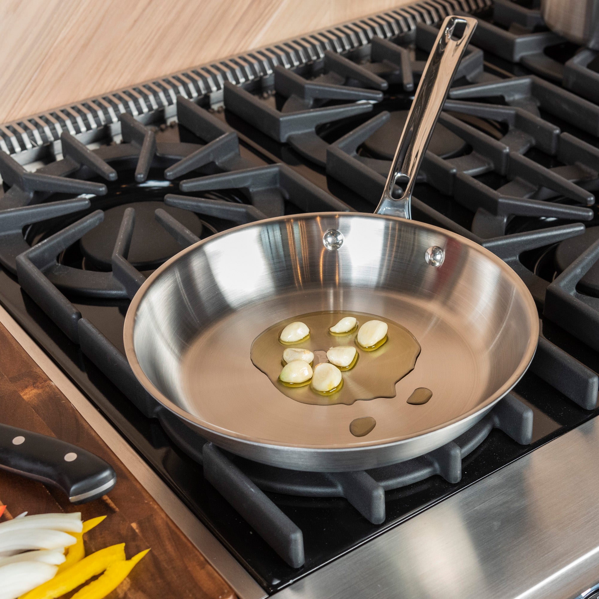 The Viking Culinary Products Viking Professional 5-Ply 10-Inch Fry Pan heats oil and garlic on a gas stove, while a knife and fresh-cut vegetables sit on the adjacent wooden counter.