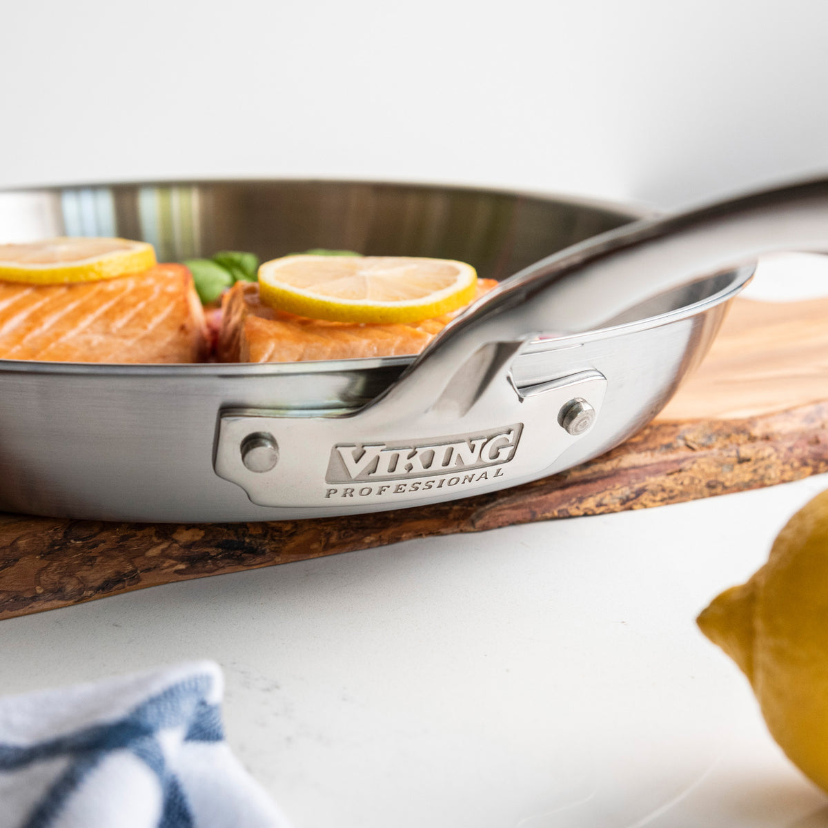 A Viking Professional 5-Ply 12-Inch Fry Pan by Viking Culinary Products, holding salmon fillets with lemon slices, rests on a wooden board. A lemon and towel lie in front, spotlighting this dishwasher safe cookware.