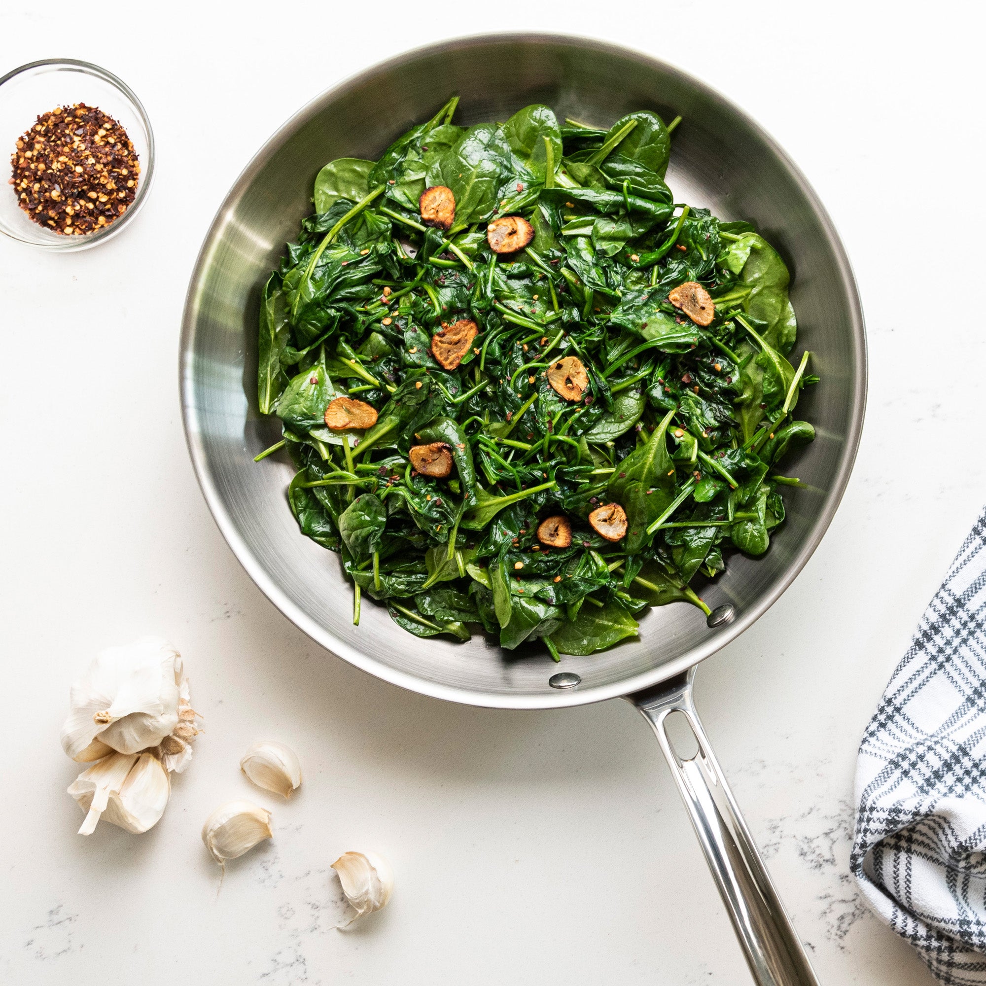 The Viking Professional 5-Ply 12-Inch Fry Pan by Viking Culinary Products, filled with spinach and garlic, sits on a white surface near red pepper flakes, garlic cloves, and a blue checkered towel. Dishwasher safe.